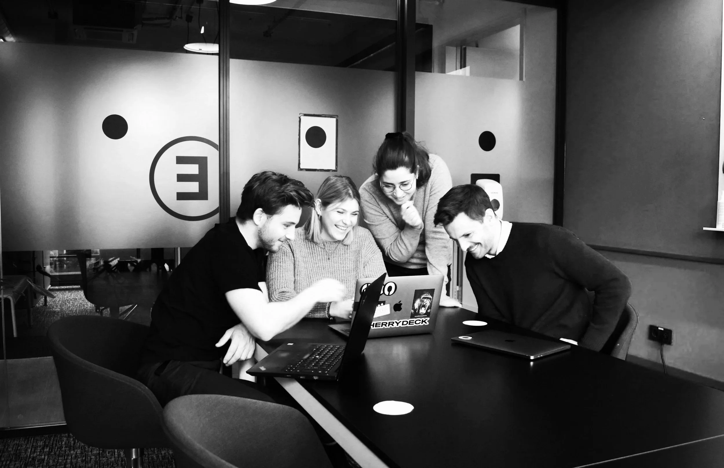 A group of five creatives gathered around a table in an office, looking at a laptop and smiling. The background features partitions with abstract logo designs.