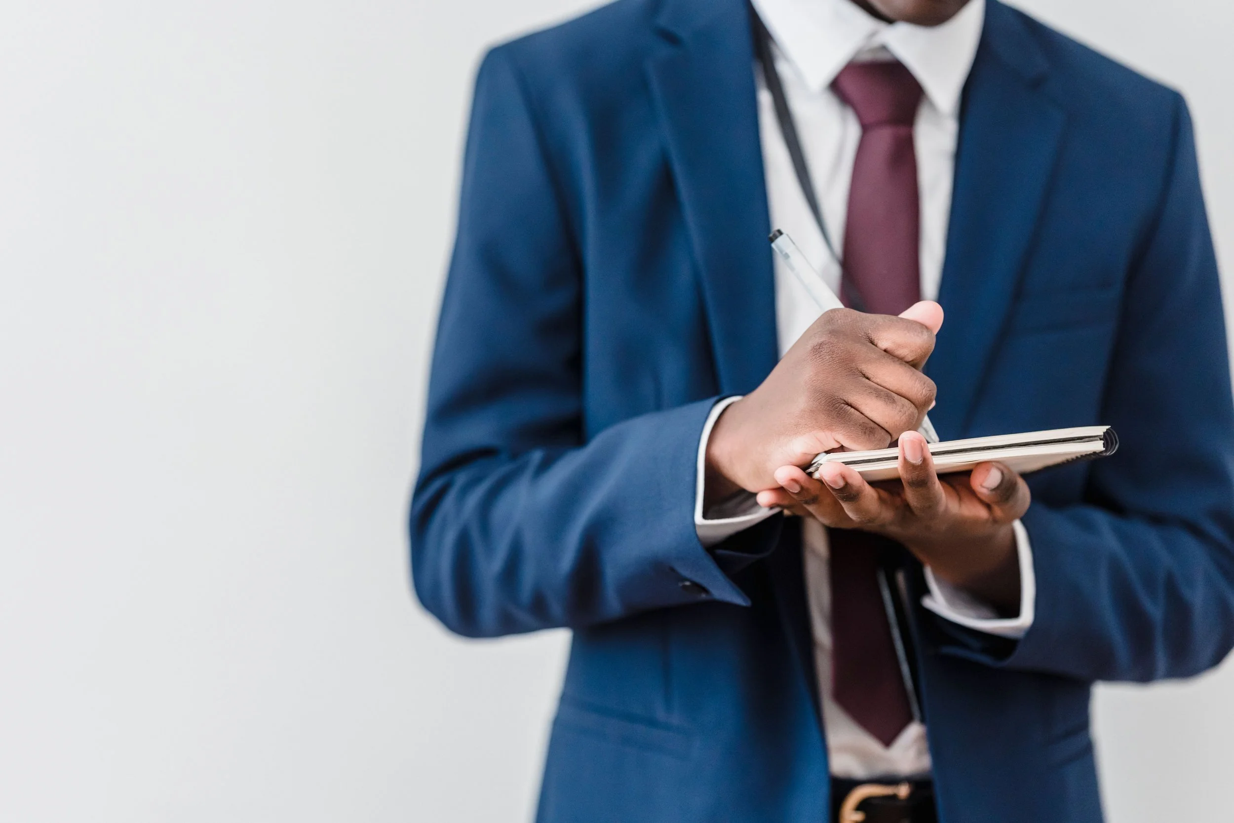 A person in a blue business suit with a maroon tie holding a pen and notepad.