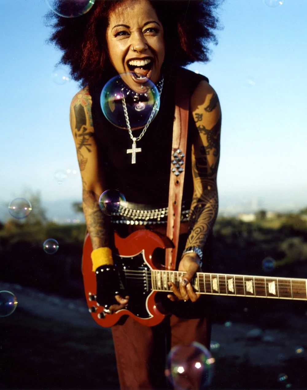 A woman with curly hair playing a red electric guitar outdoors, smiling broadly, with bubbles floating around her, wearing a black tank top and a silver cross necklace.