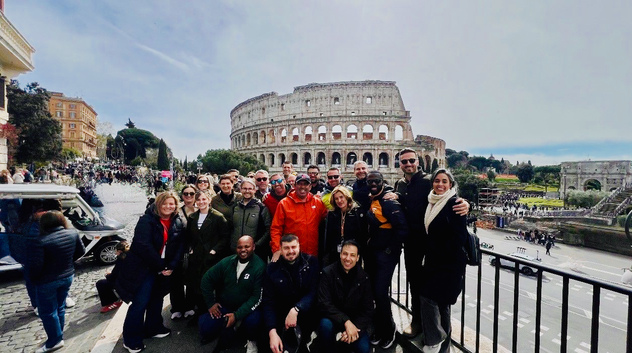 Tour group standing in front of the Colosseum in Rome, Italy, with other tourists, cars, and historic ruins in the background.