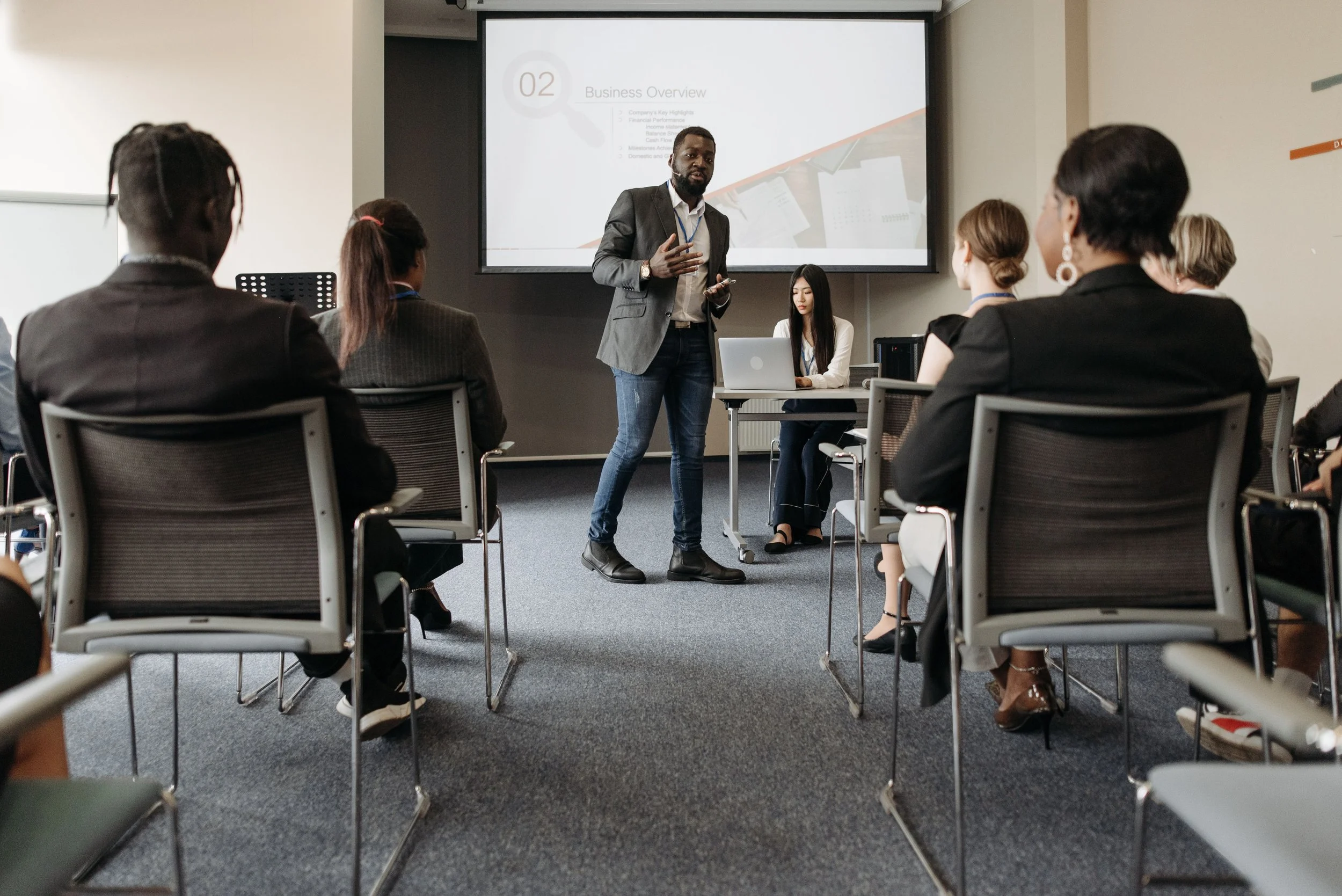 A man in a suit giving a presentation to a group of professionals in a conference room. The group is seated and listening attentively, with a large screen displaying a business overview in the background.