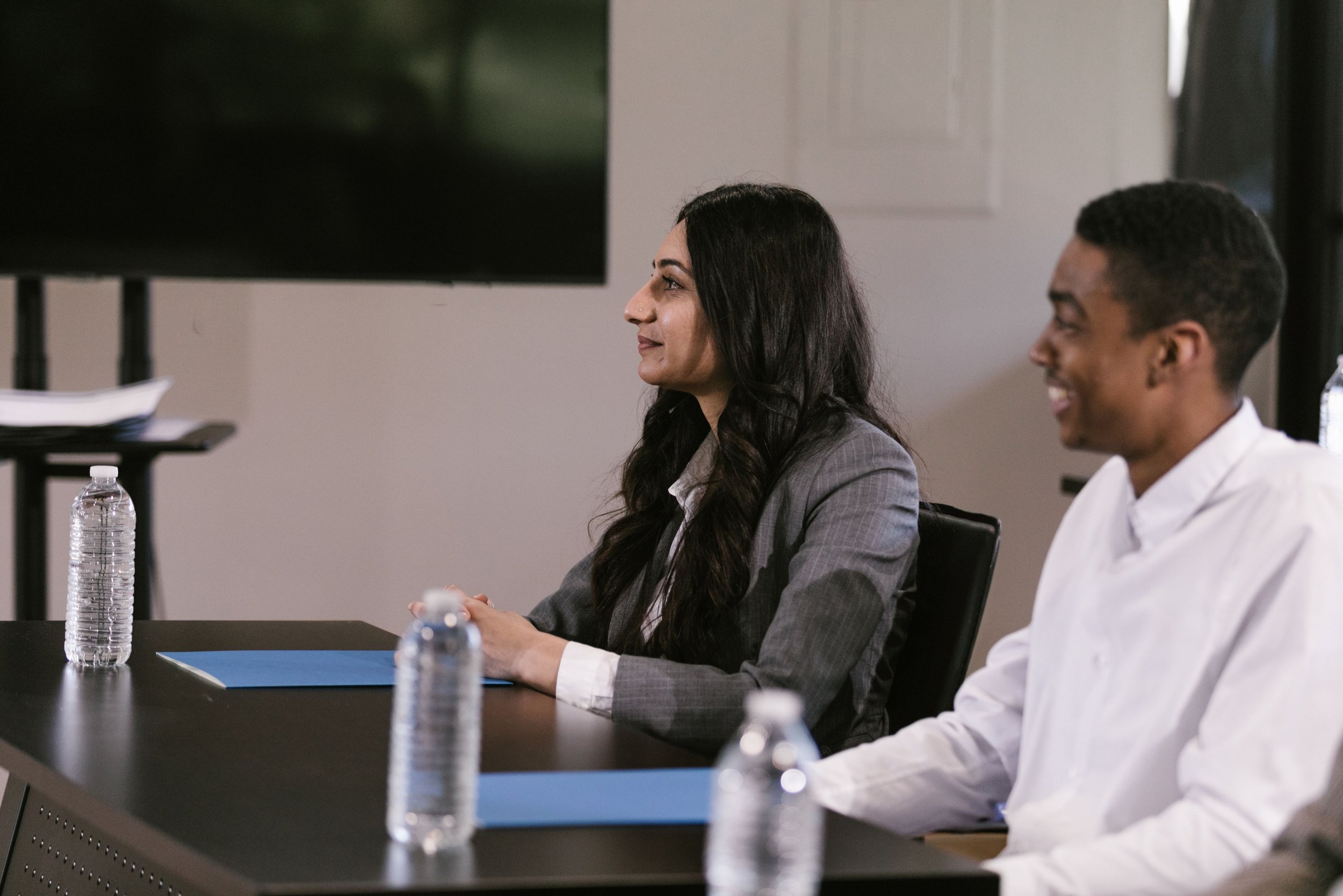Two people sitting at a conference table during a formal meeting, with water bottles on the table.