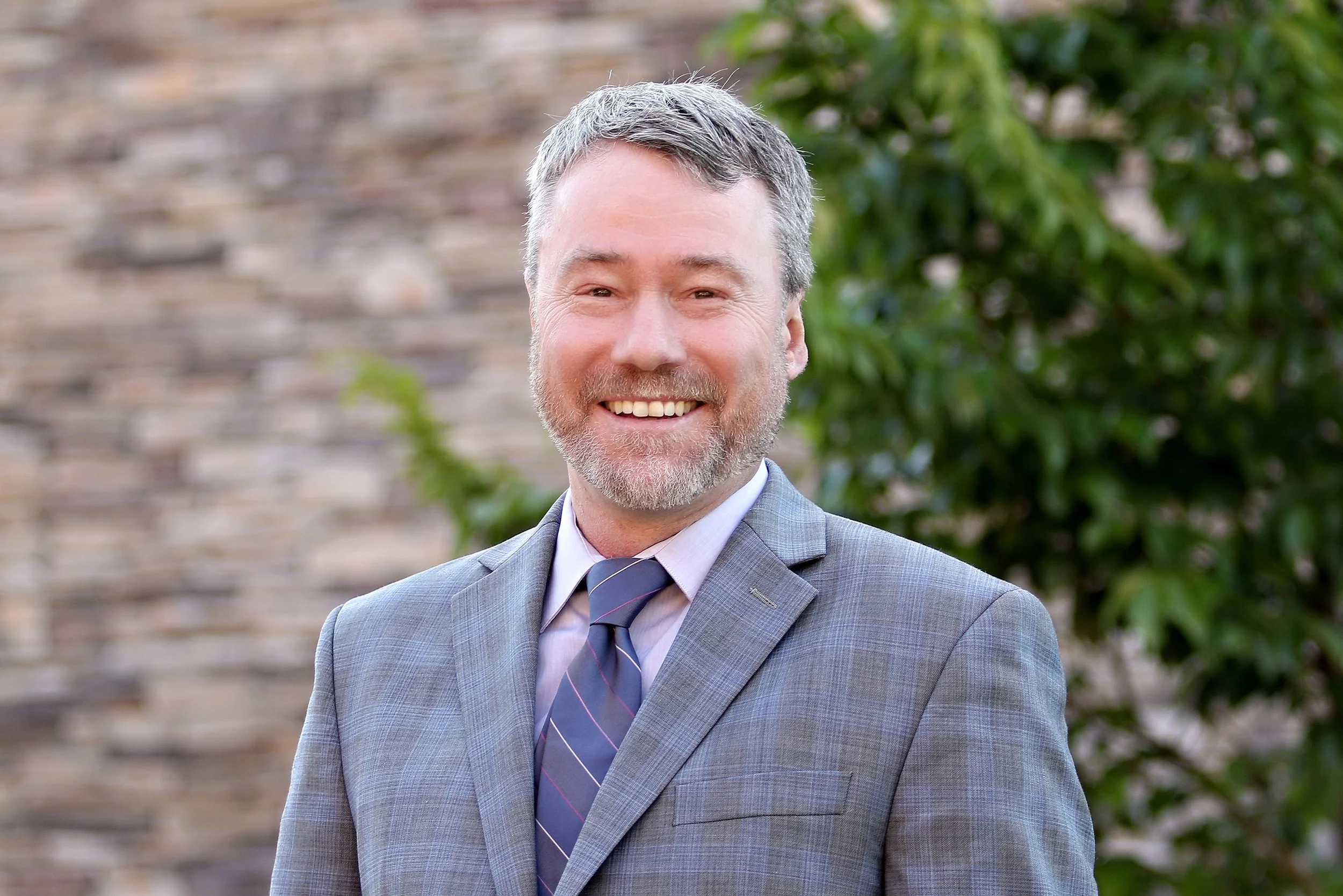 A middle-aged man with short gray hair, beard, and mustache smiling outdoors, wearing a gray suit, white shirt, and a striped tie, with a background of a stone wall and green leafy foliage.