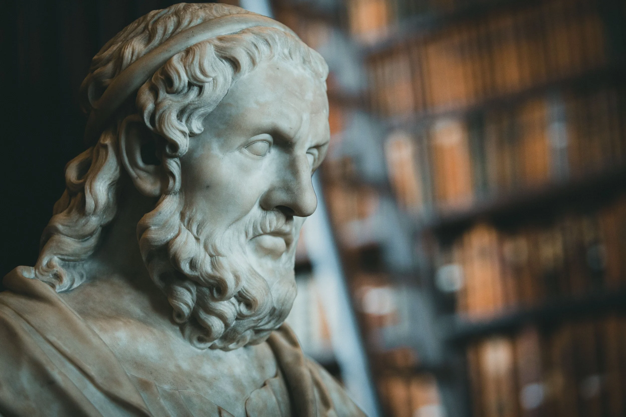 Close-up of a marble bust sculpture of a man with a beard and flowing hair, set against a background of bookshelves filled with books.