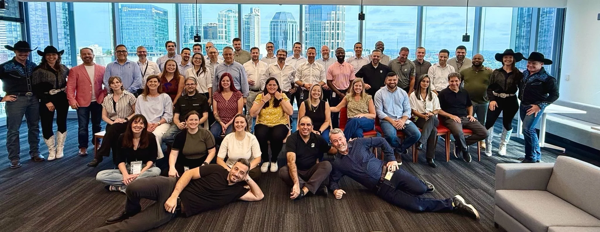 Group photo of diverse employees in a high-rise office with city skyline views, some dressed in cowboy attire, smiling and sitting or standing together.