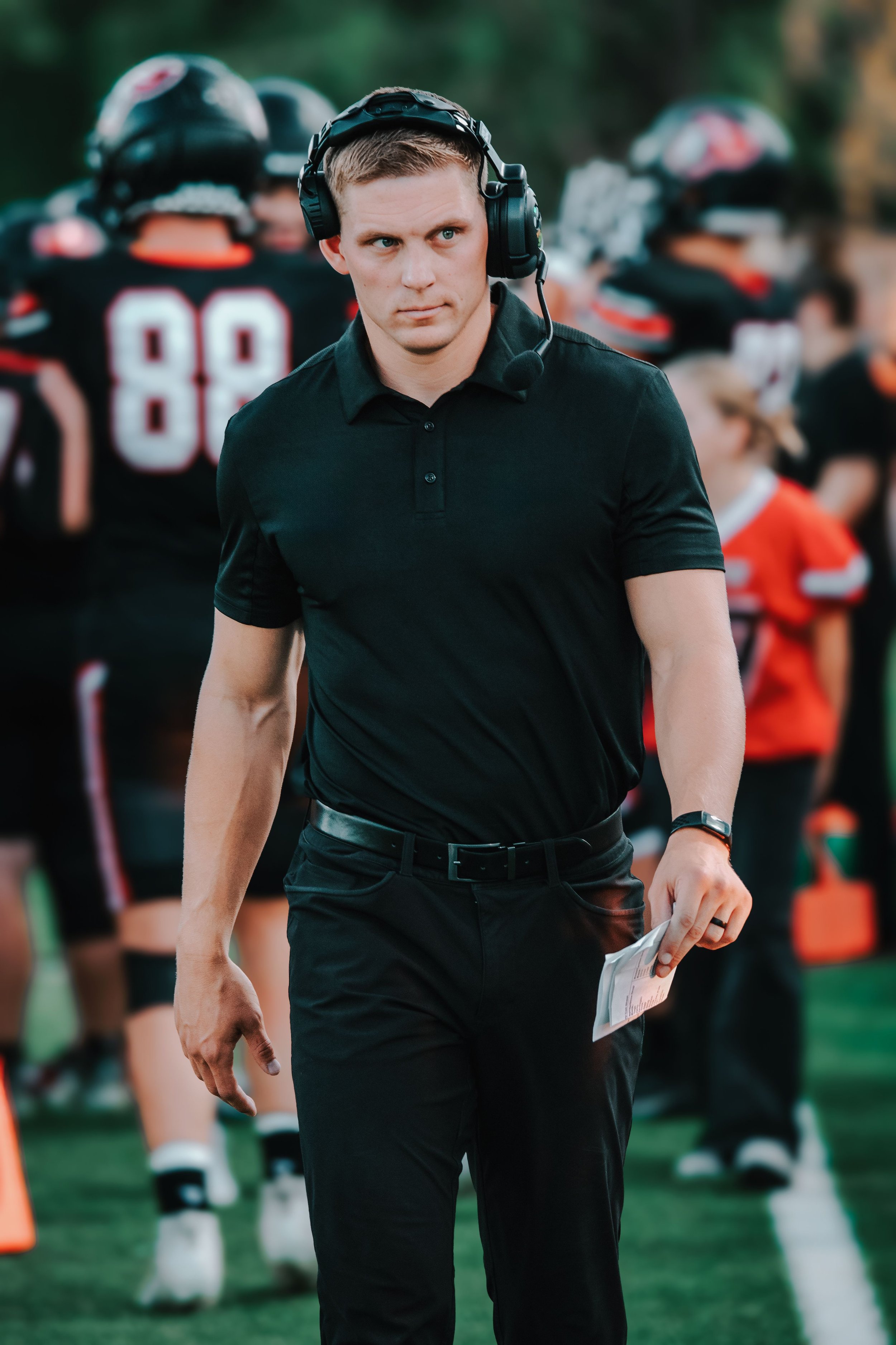 A man in a black polo shirt and black pants, wearing a headset with a microphone, holding a piece of paper, standing in front of football players on the sidelines.