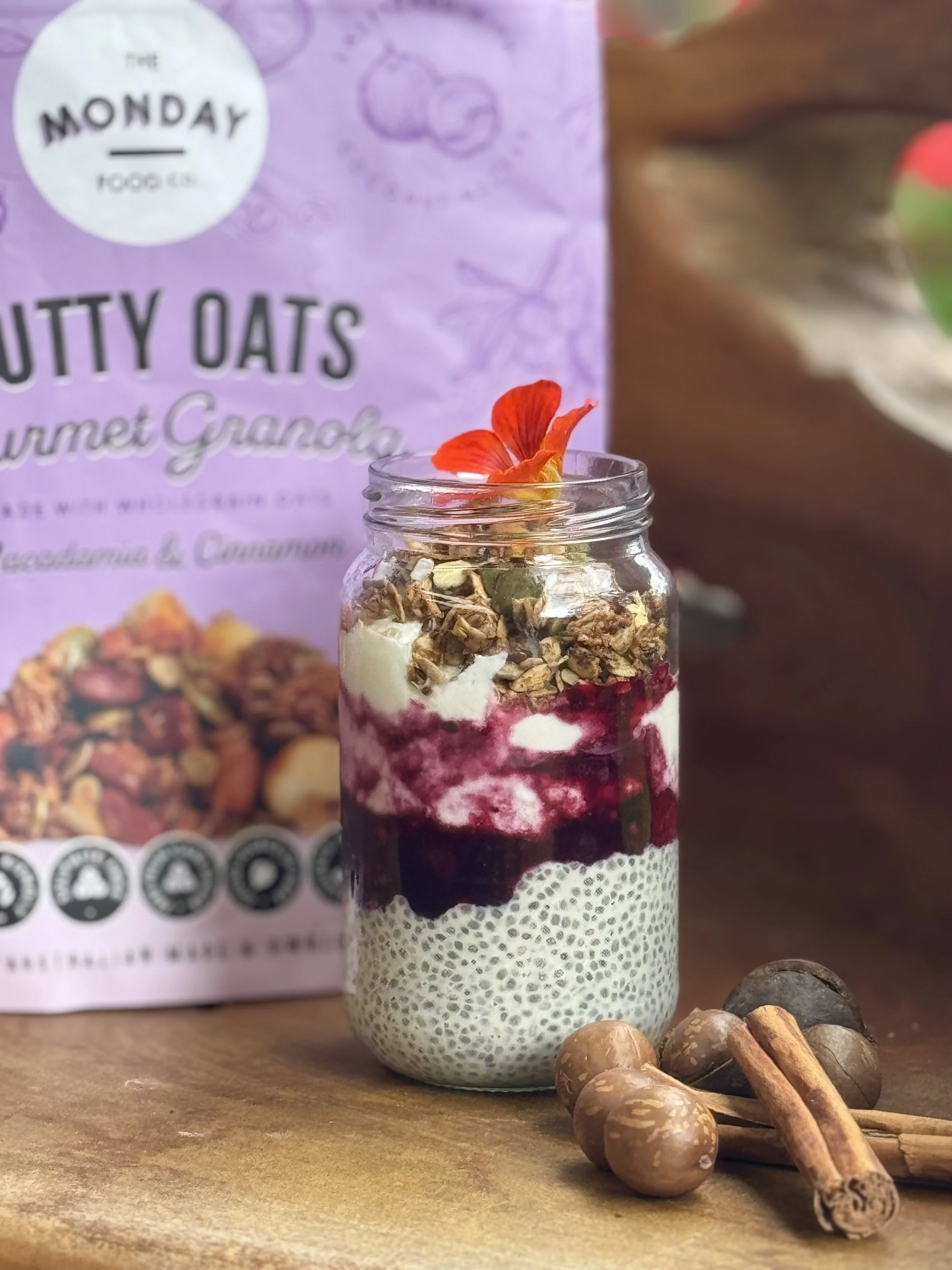 A jar filled with layered granola, yogurt, and berries, topped with an edible flower. Behind the jar, there is a purple snack package with the text 'Nutty Oats' and some oats. Several hazelnuts and cinnamon sticks are placed on the wooden surface around the jar.
