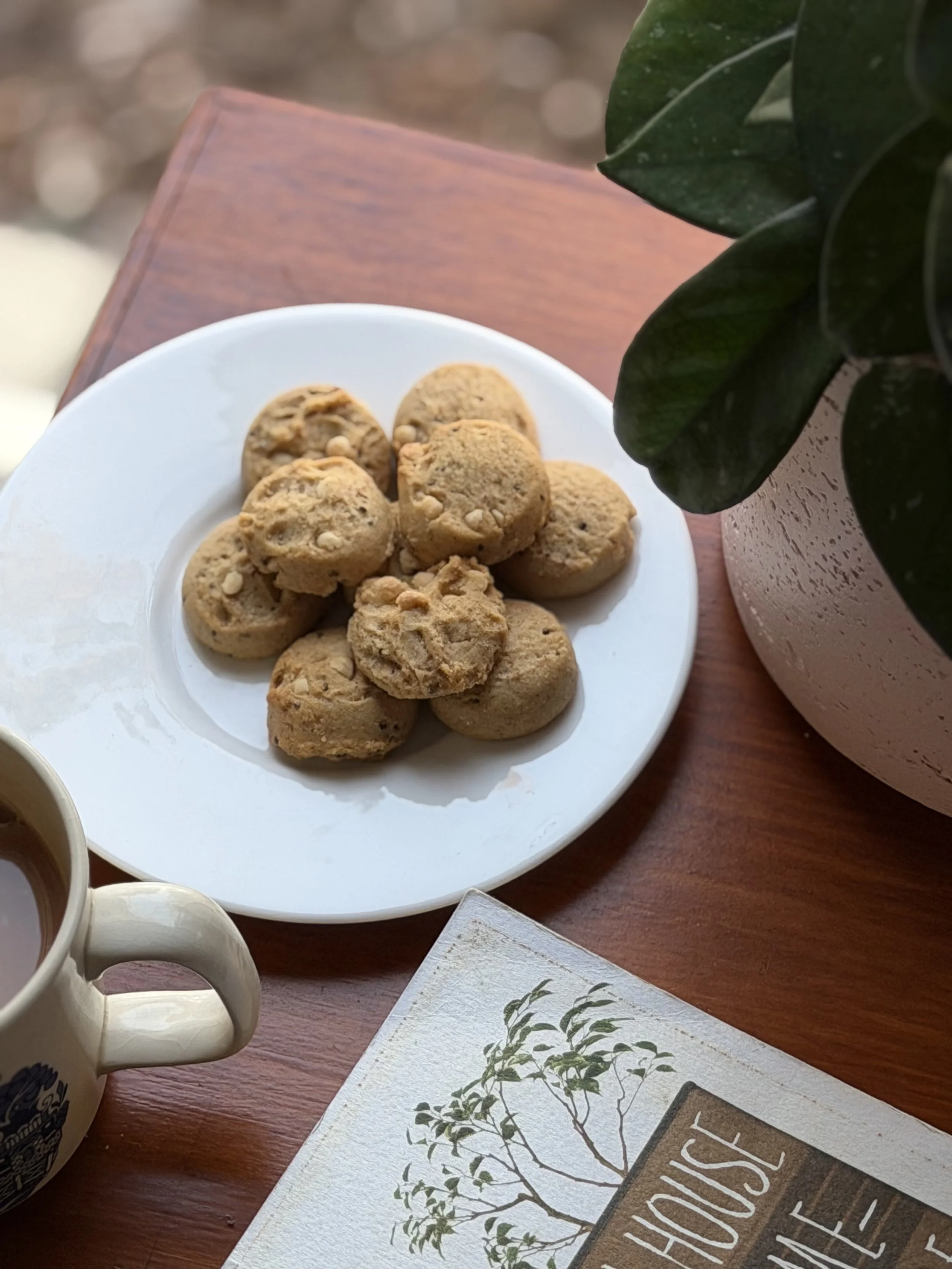 A white plate with oatmeal cookies on a wooden table. To the right is a plant with large green leaves in a pink pot. Part of a mug and a card with the word 'HOUSE' visible are also in the image.