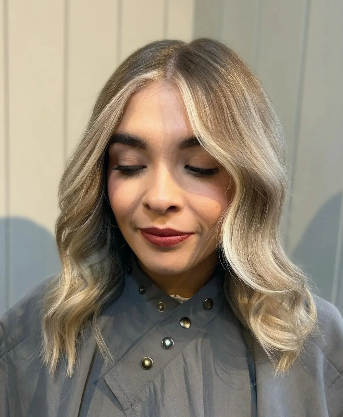 A woman with long wavy blonde hair, closed eyes, and a slight smile. She is wearing a gray top with metallic button details, against a neutral background.