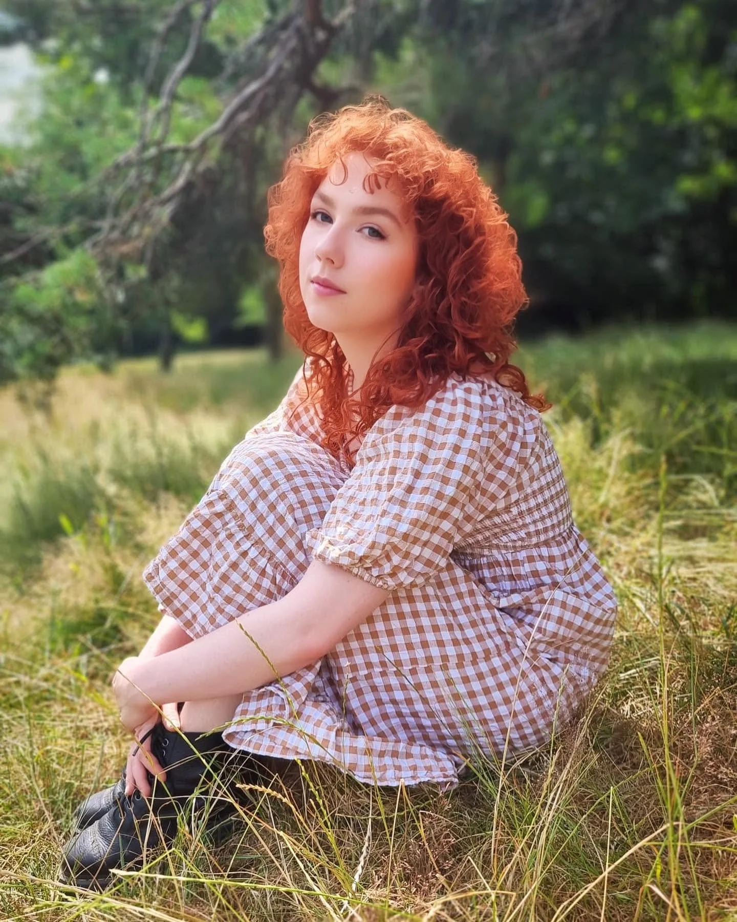 A young woman with curly red hair and fair skin sitting in a field of grass with trees in the background.