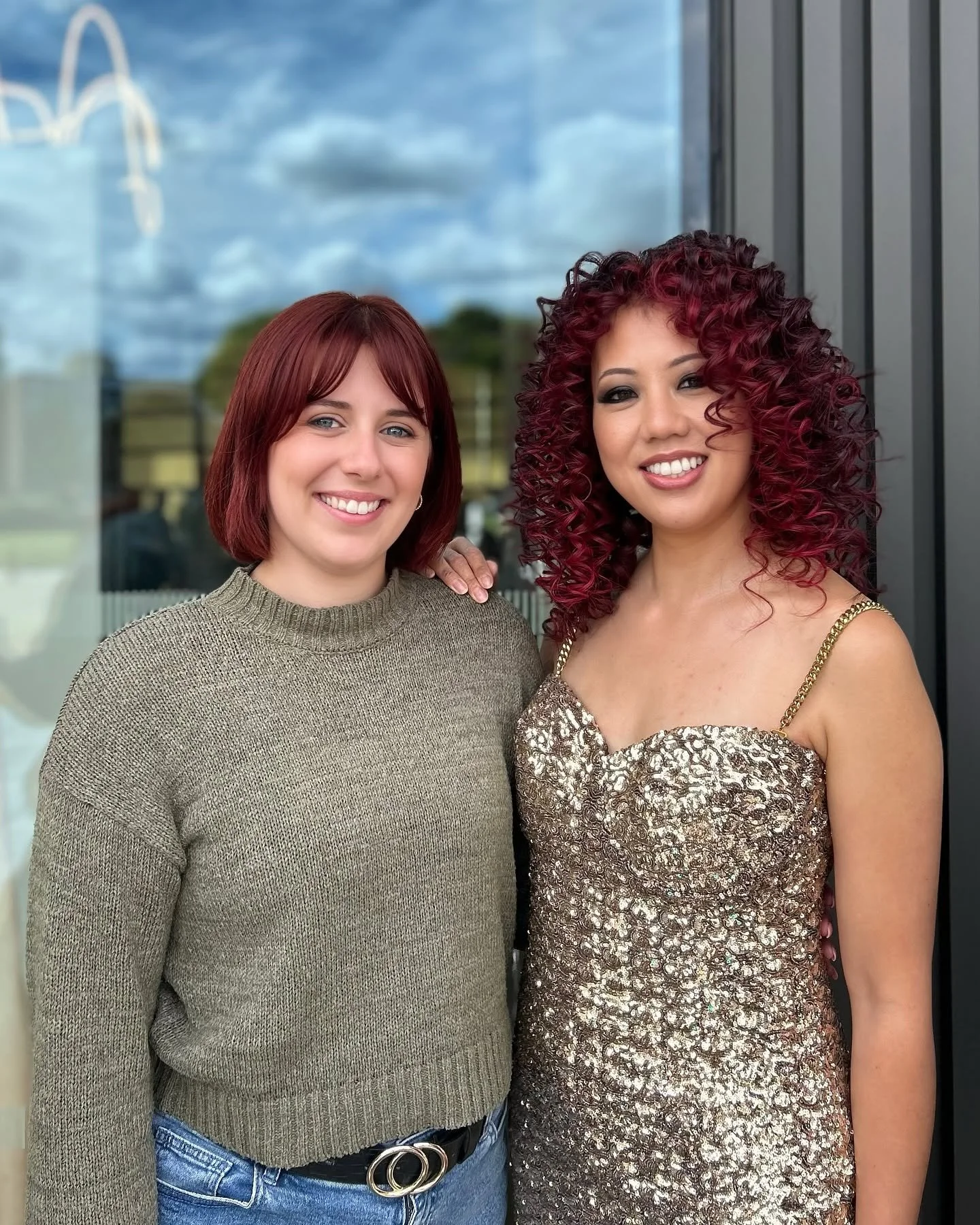 Two smiling women standing side by side outdoors in front of a glass window, with a reflection of the sky and trees, one in a brown sweater and blue jeans, the other in a gold sequined dress with curly red hair.