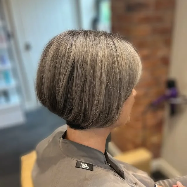 Side view of an older woman with a sleek, chin-length gray bob haircut sitting in a salon chair.