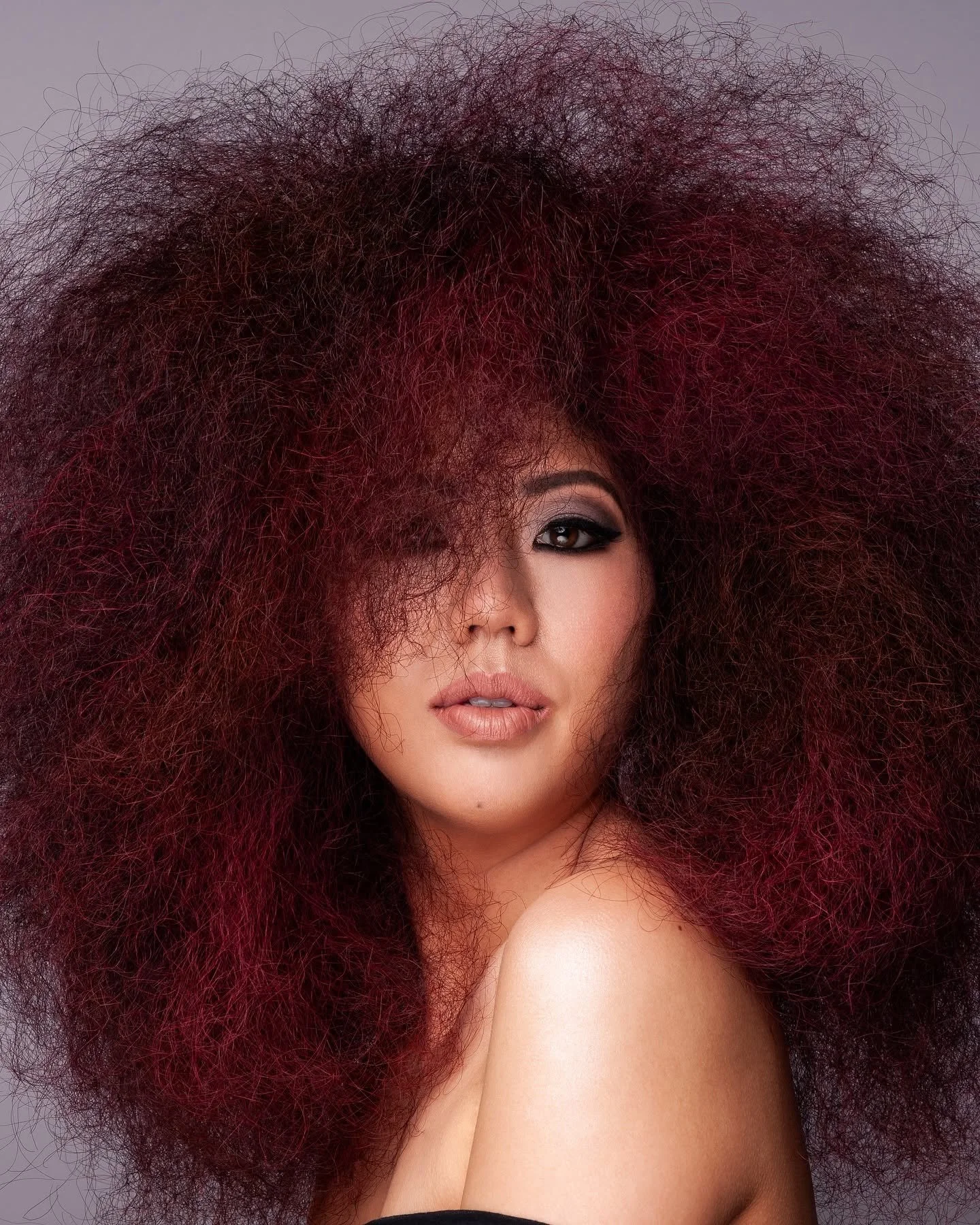 A woman with voluminous, curly hair that is dark red at the roots and fades to magenta at the tips, partially covering one eye.