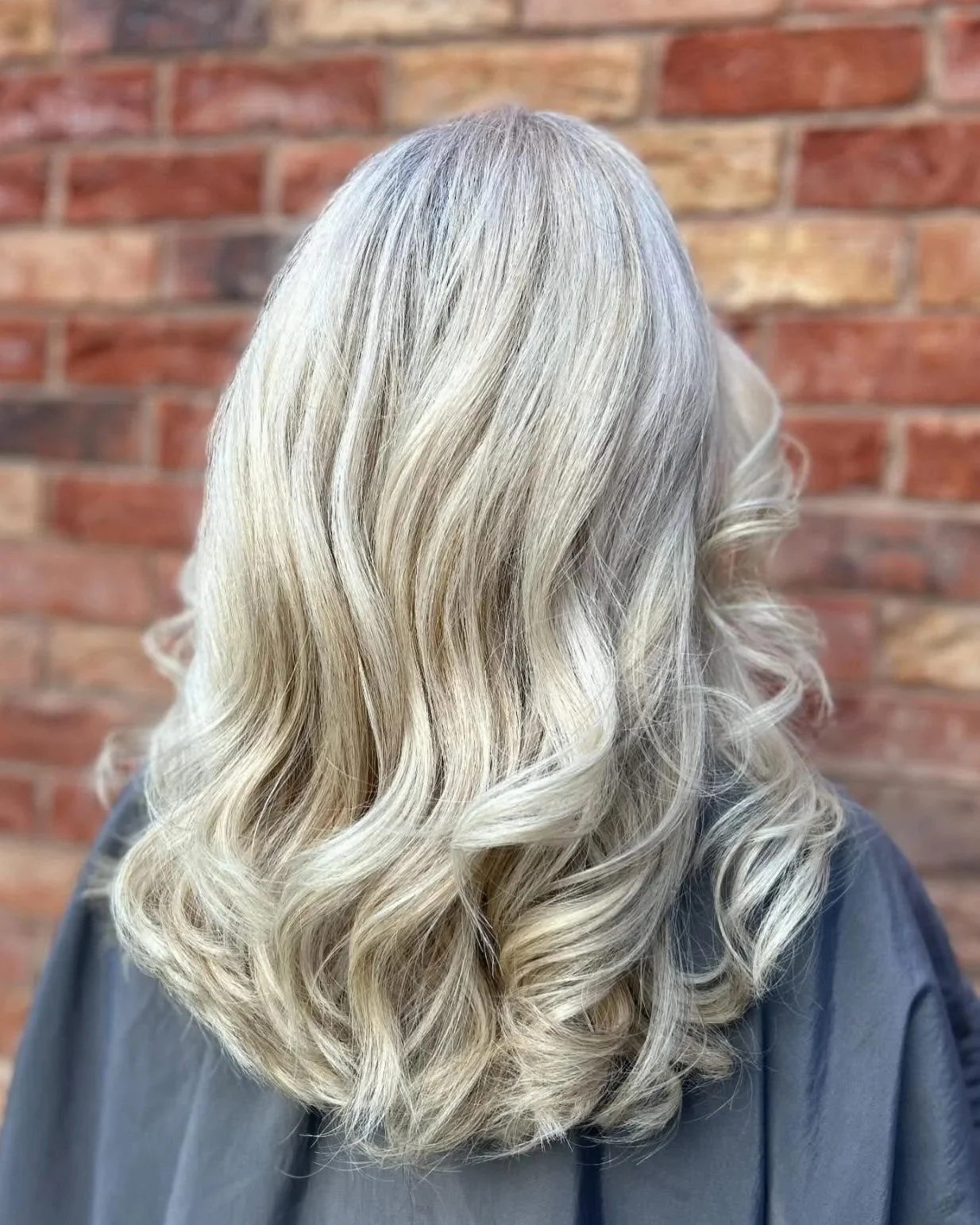 Back view of a woman with long, wavy, platinum blonde hair with highlights, standing in front of a red brick wall.