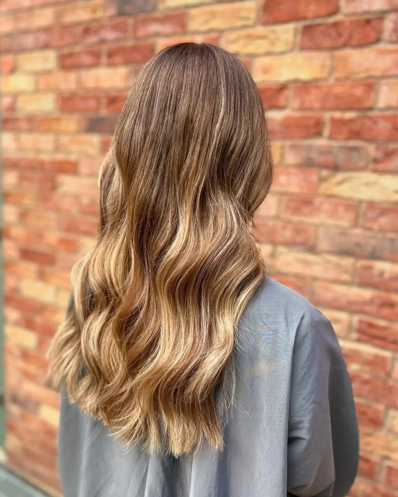 Back view of a woman with long, wavy, highlighted hair standing against a brick wall.