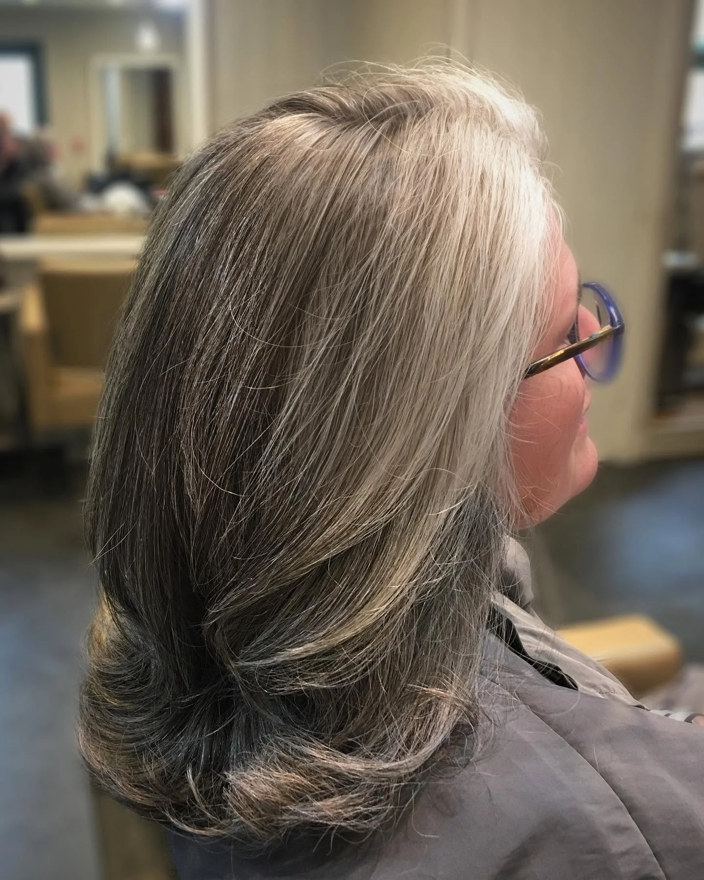 Side view of an older woman with shoulder-length gray hair, sitting in a salon or similar setting, wearing glasses and a gray cape.