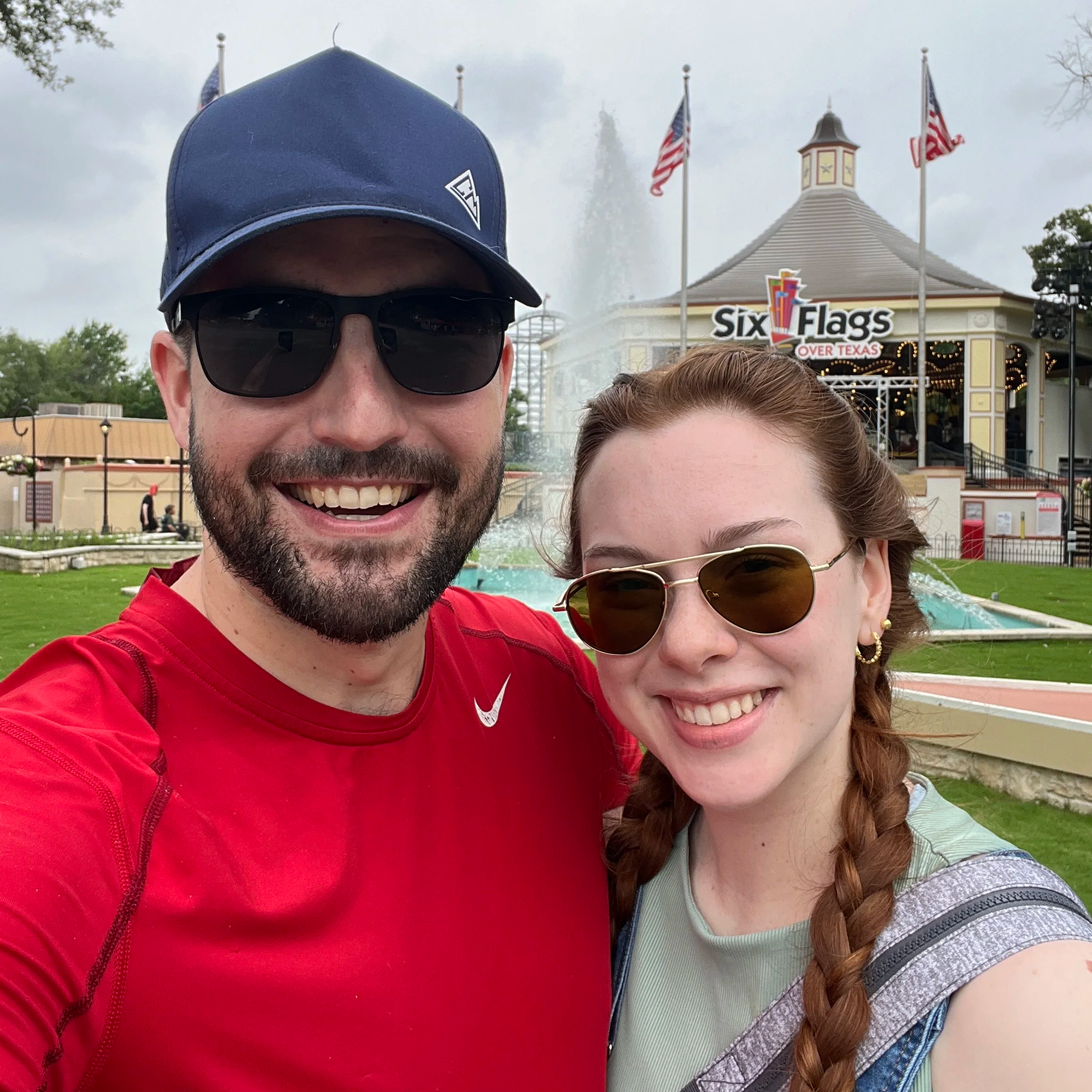A smiling man wearing sunglasses and a navy blue cap standing next to a woman with long braided hair, both wearing casual clothes at Six Flags amusement park in Over Texas, with the park's entrance building and American flags in the background.