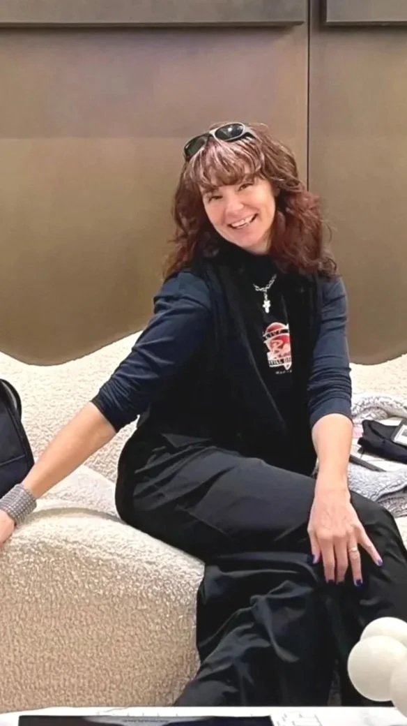 A woman with curly brown hair wearing sunglasses on her head and a black shirt, sitting on a beige sofa, smiling, with a dark background.