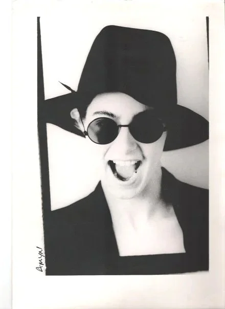 Black and white photo of a person wearing a wide-brimmed hat and sunglasses, smiling with their mouth open.