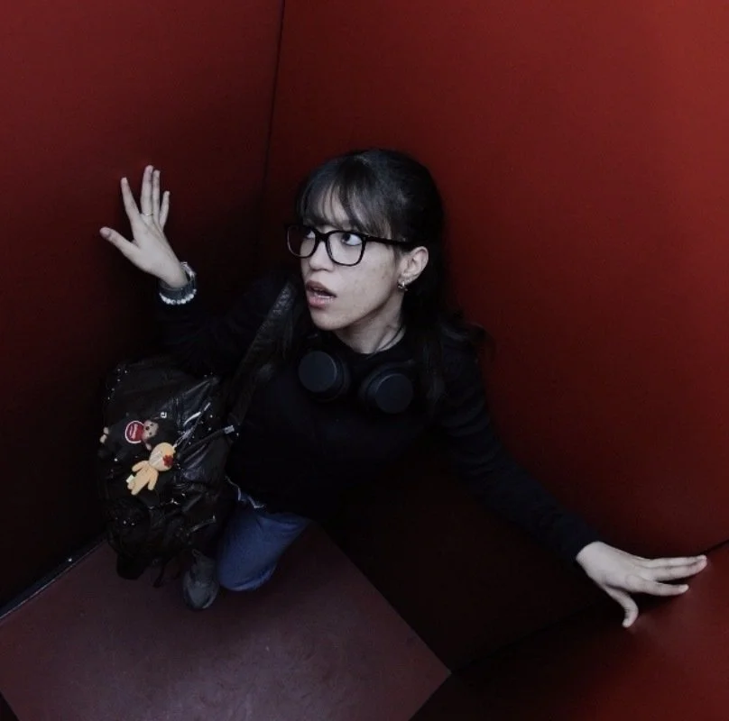 A woman with glasses and headphones around her neck crouches in a small red-walled space, looking up with a surprised or confused expression.