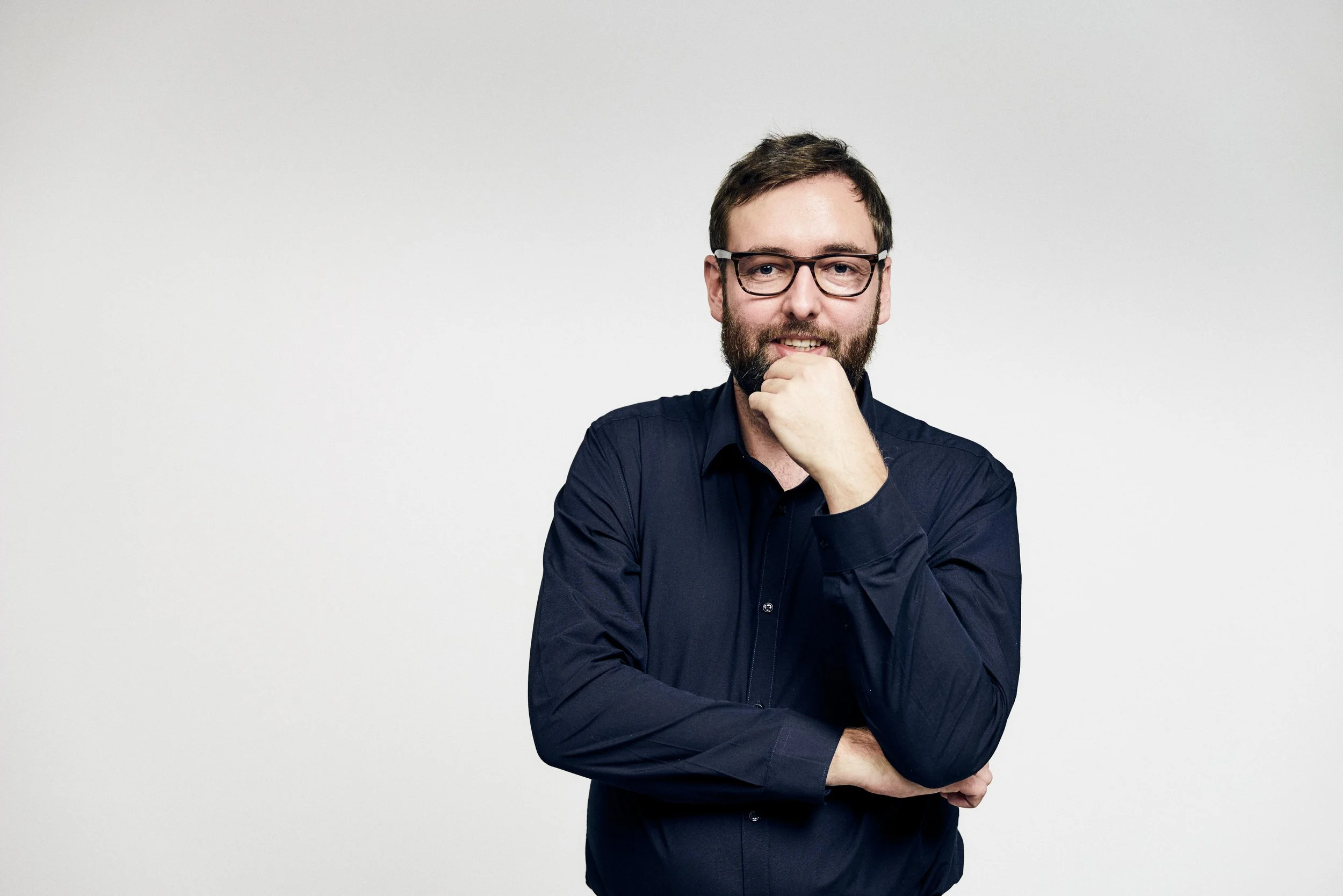 Ein Mann mit Glatze, Vollbart, Brille und dunklem Hemd steht vor einem weißen Hintergrund. Er hat die Hand vor den Mund und schaut nach vorne.