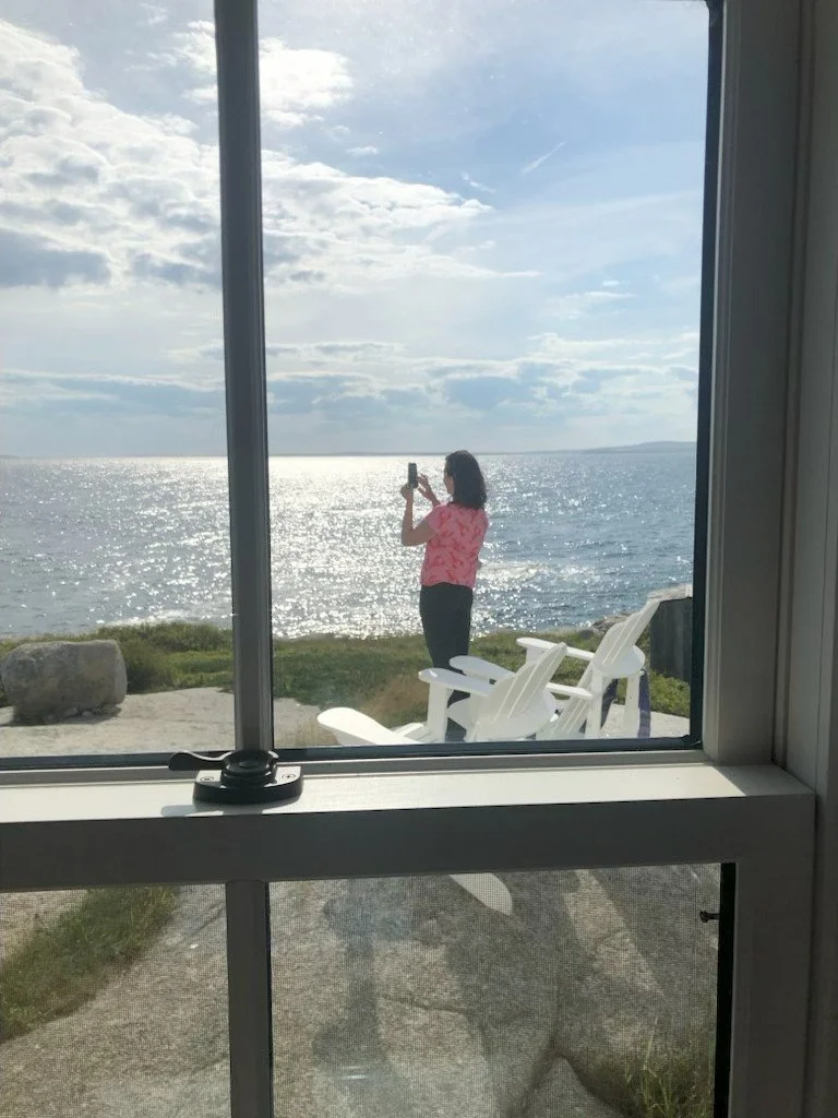 A woman in a pink shirt and dark pants stands by the water taking a photo with her phone, sitting on a chair outdoors near the ocean, viewed through a window.