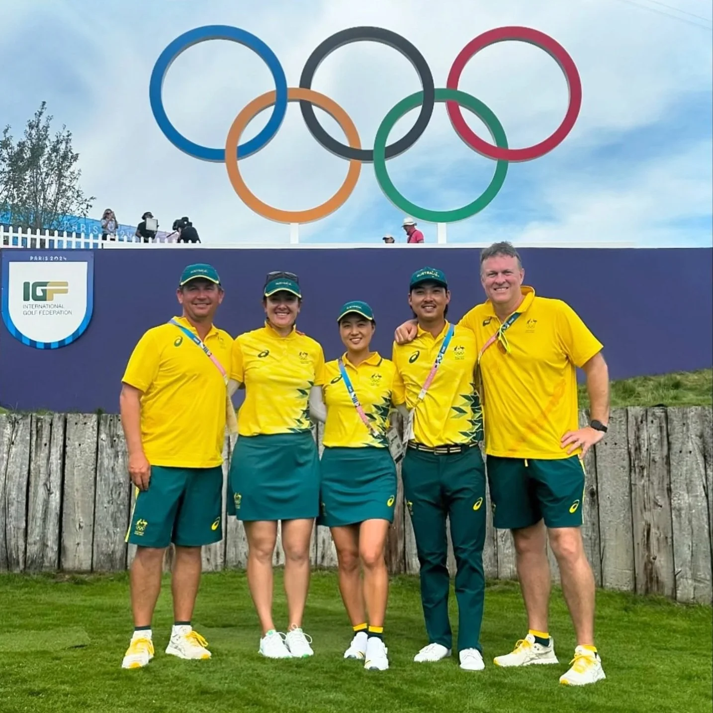 Five Australian athletes in yellow and green uniforms standing together in front of a fence, with the Olympic rings logo and a purple sign with the International Golf Federation logo behind them.