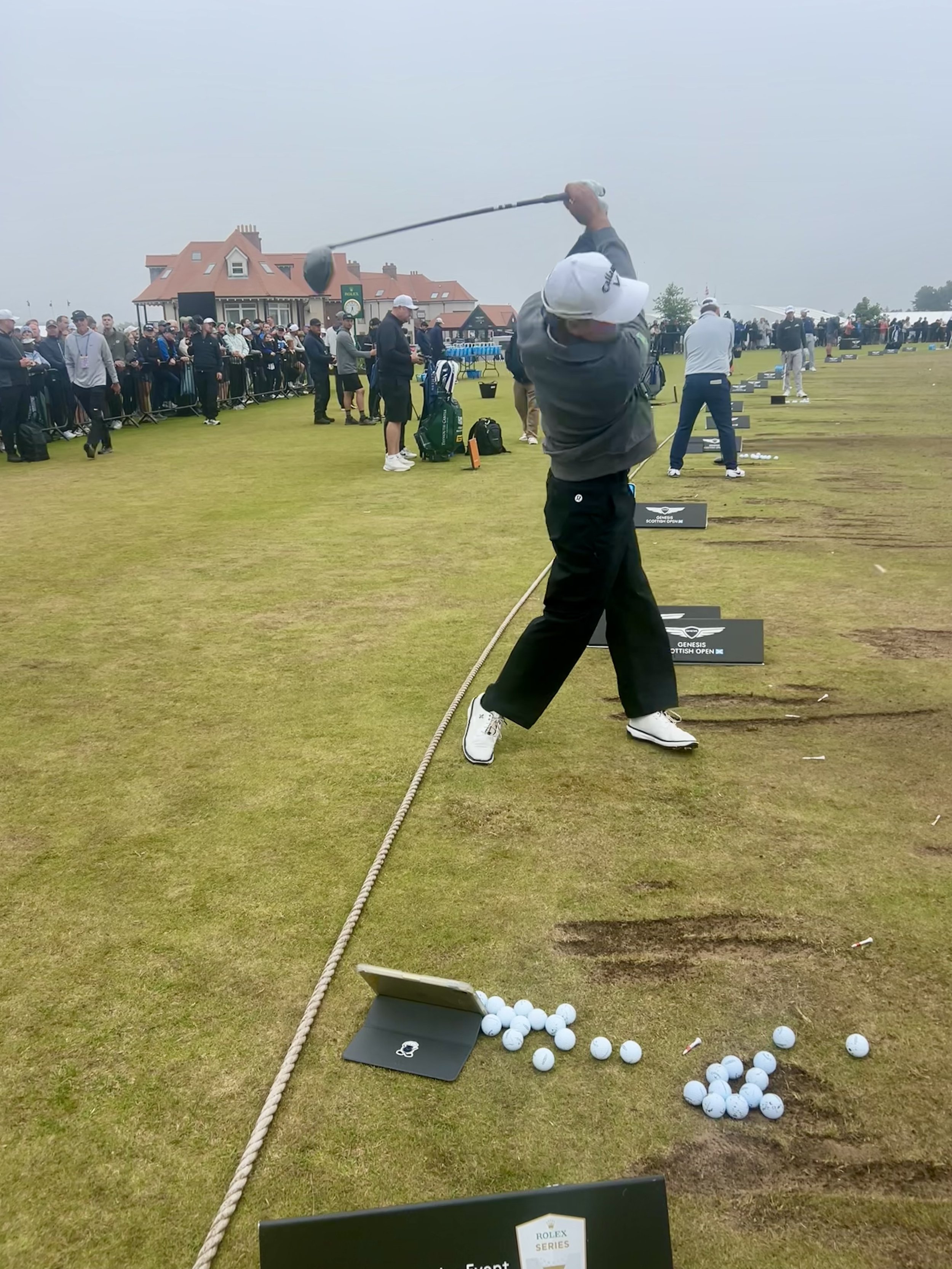 A golfer swings a club at a driving range during a golf tournament, with many spectators observing in the background.