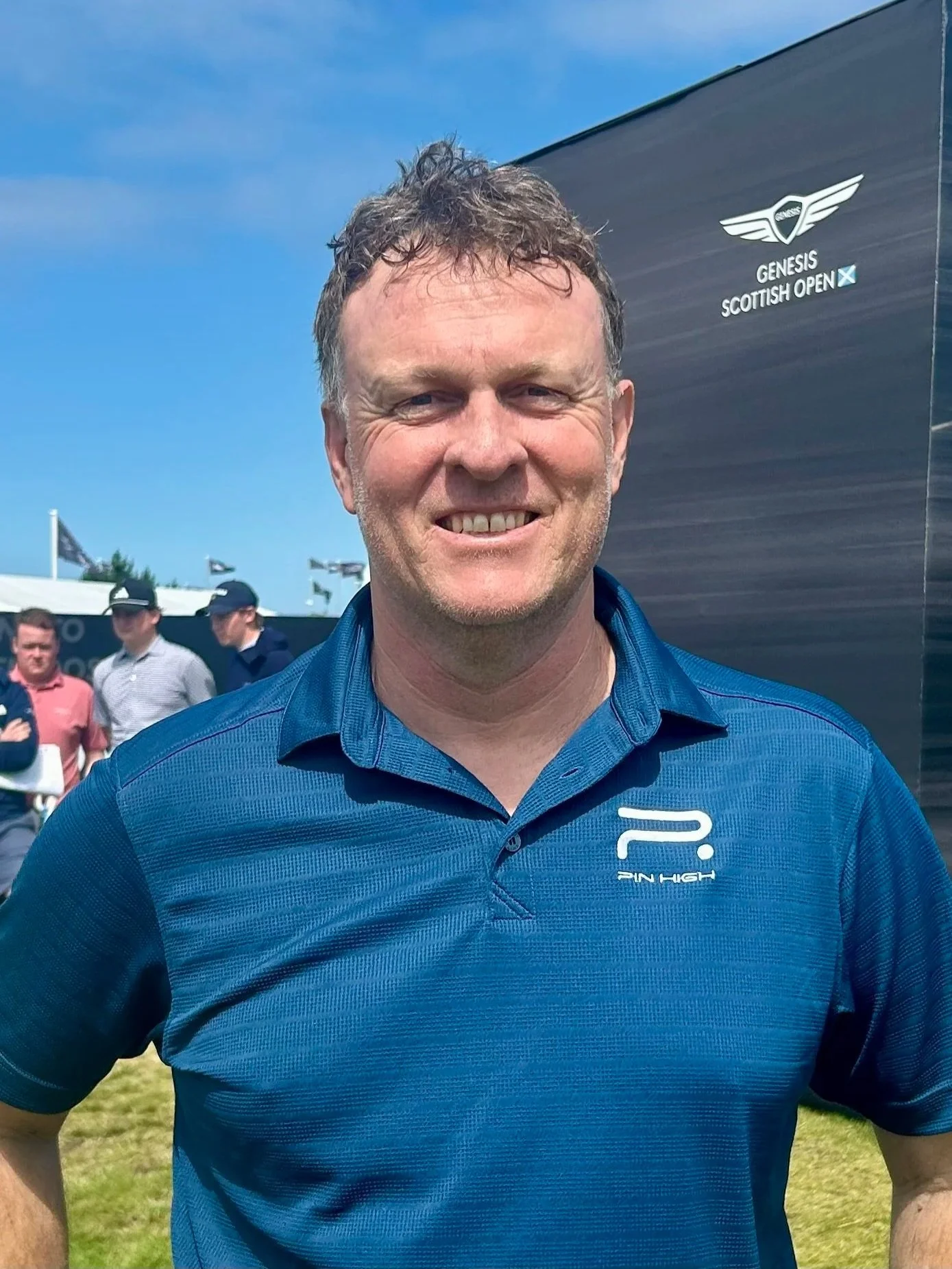 Close-up of a man with short, curly brown hair, smiling, wearing a blue polo shirt with a logo that says 'PIN HIGH,' standing outdoors at the Genesis Scottish Open golf tournament. There is a black tent with the Genesis logo and a white glider emblem behind him, and several people in the background.