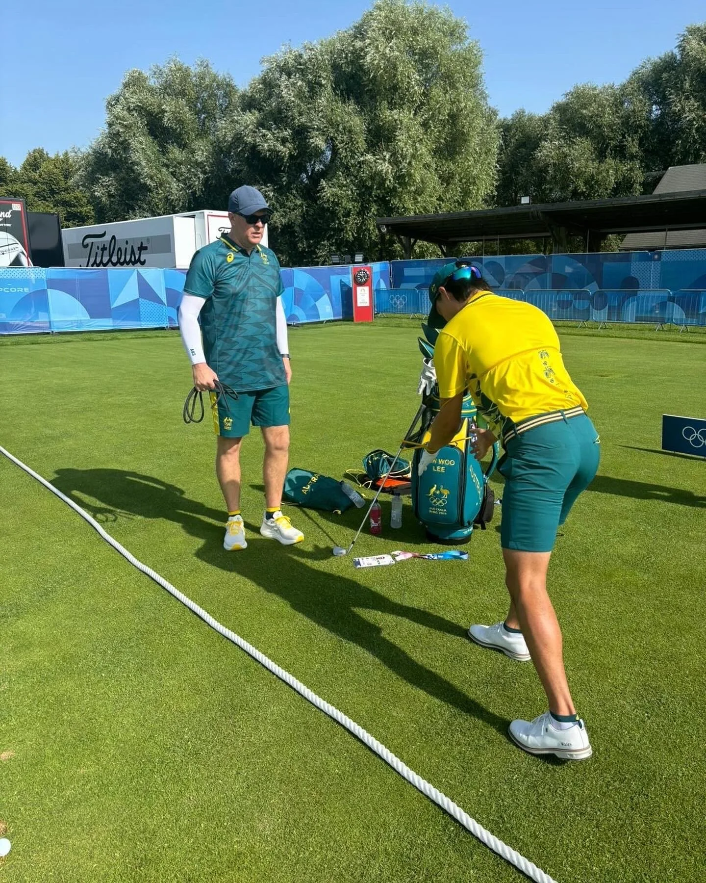 A golfer wearing a yellow shirt and teal shorts is preparing to hit a shot on a green golf course, with a male caddy or coach standing nearby.