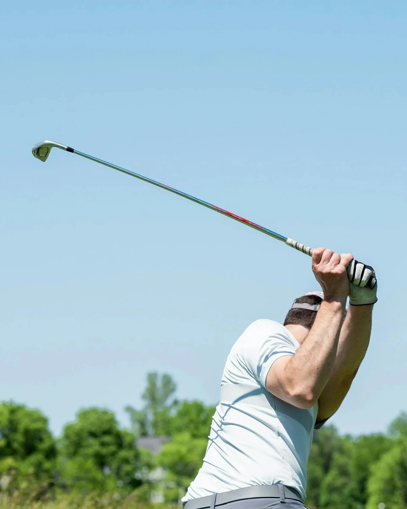 A male golfer in a white shirt and cap is swinging a golf club on a golf course with a clear blue sky and green trees in the background.