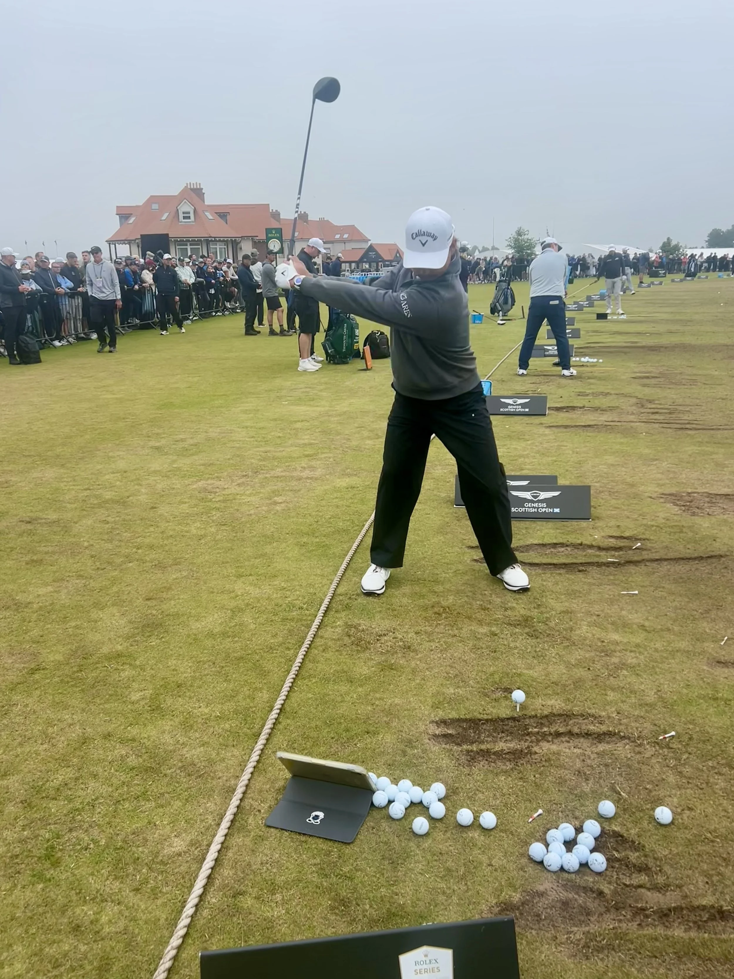 A golfer practicing his swing at a driving range during a golf tournament, with many spectators watching in the background.