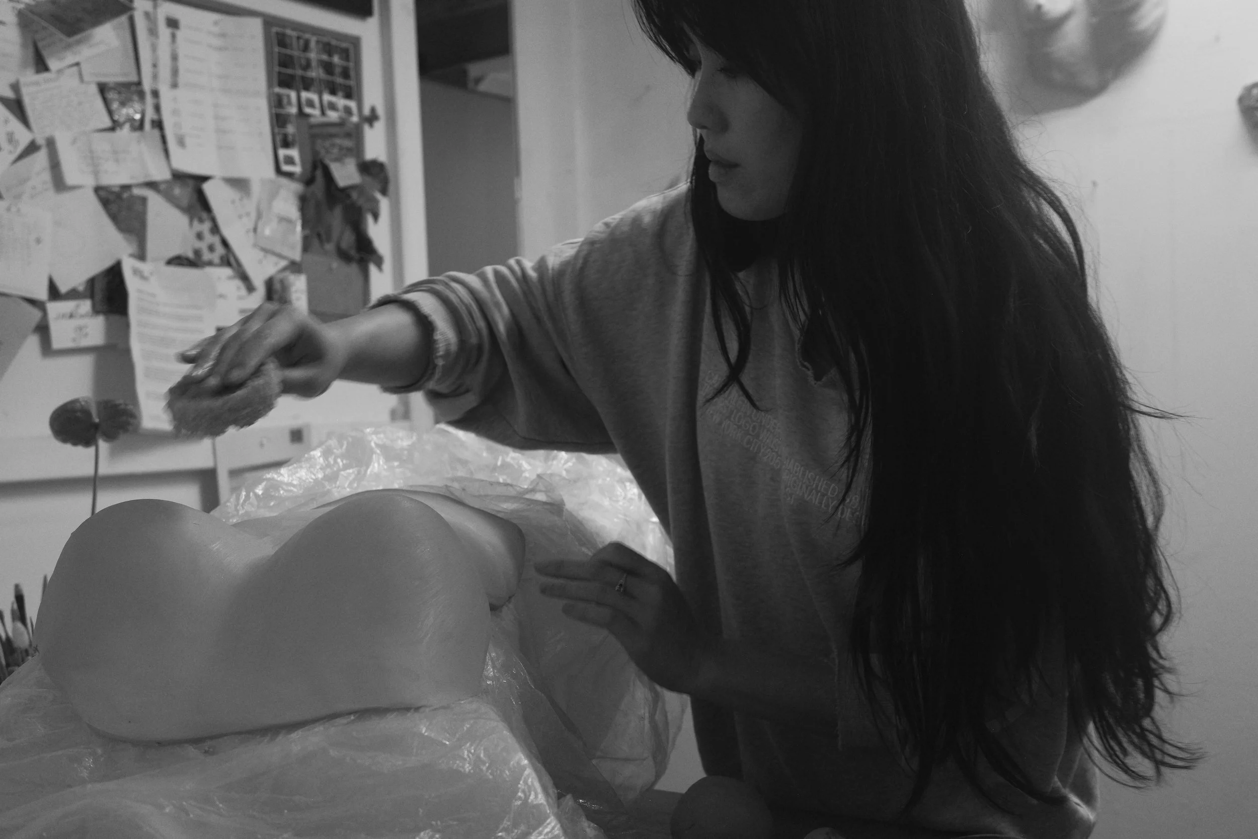 A woman with long dark hair is reaching for an object on a table, with a busy corkboard filled with papers and photos in the background.