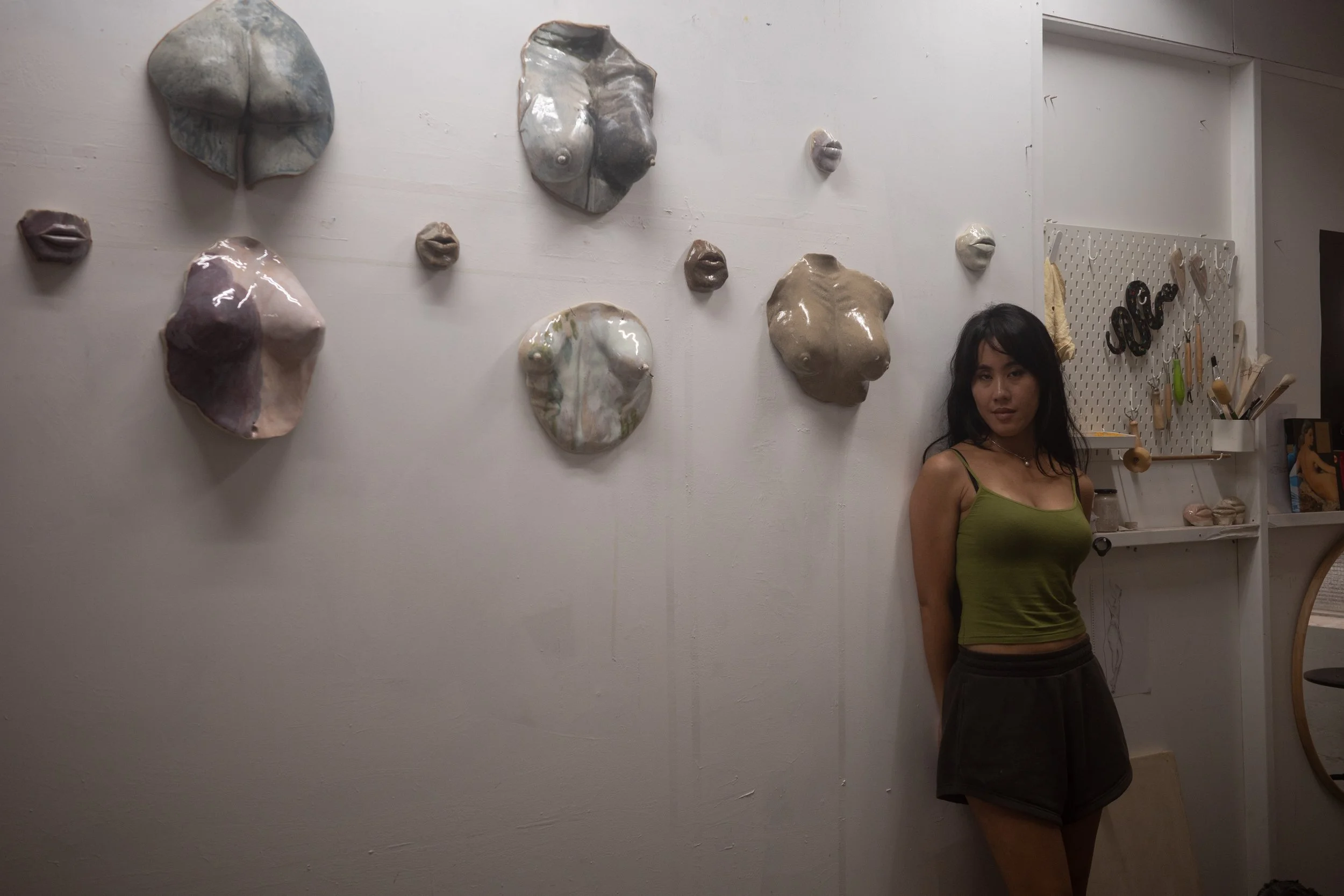 A woman with dark hair in a green tank top and black shorts standing against a white wall adorned with ceramic sculptures of lips and abstract shapes. A pegboard with various tools is visible to the right.