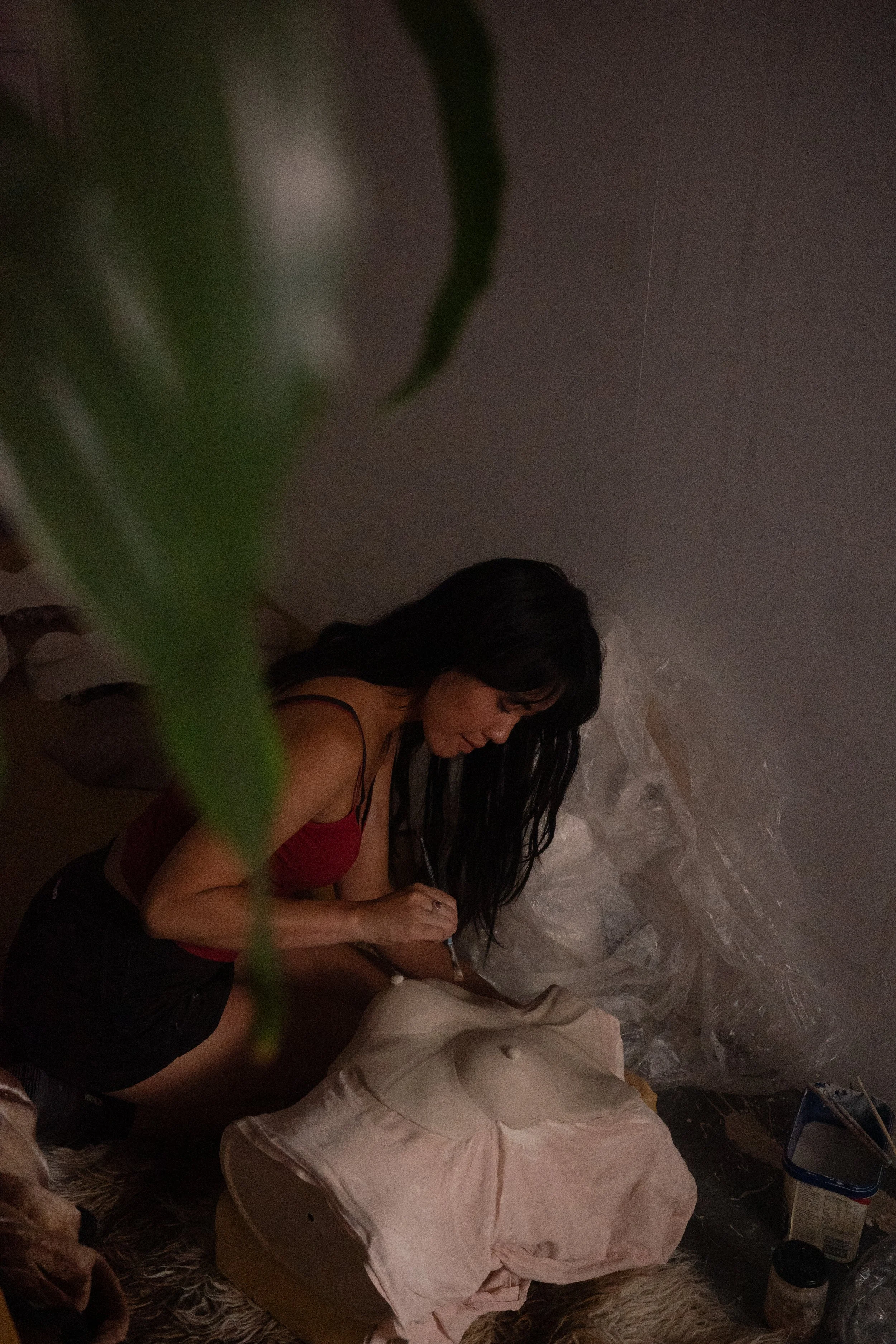 A woman with long dark hair, wearing a red top, sitting on the floor. She is focused on an activity involving fabric or clothing, with various materials around her. The setting appears to be indoors, with a textured wall in the background. A leafy plant partially obscures the view in the foreground.