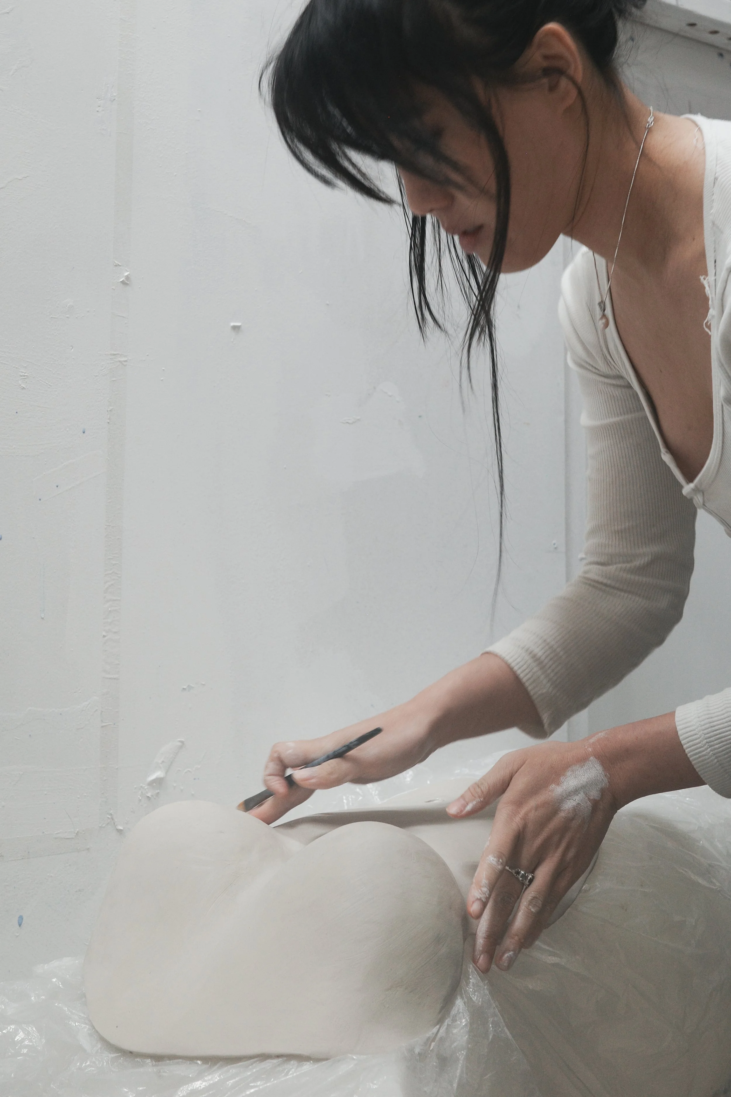 Person working on a clay sculpture, shaping with a small sculpting tool, in a minimalistic studio with white walls.