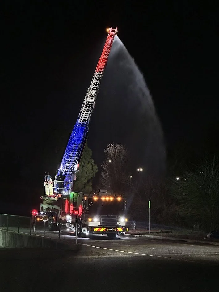 Fire truck with an extended ladder spraying water at night, illuminated by colorful lights.