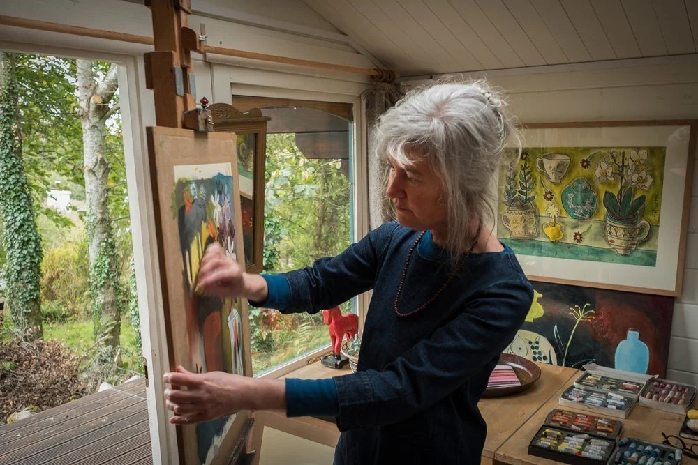 An older woman with gray hair, wearing a dark long-sleeve shirt, painting on a canvas in a room with large windows and woodland view outside.