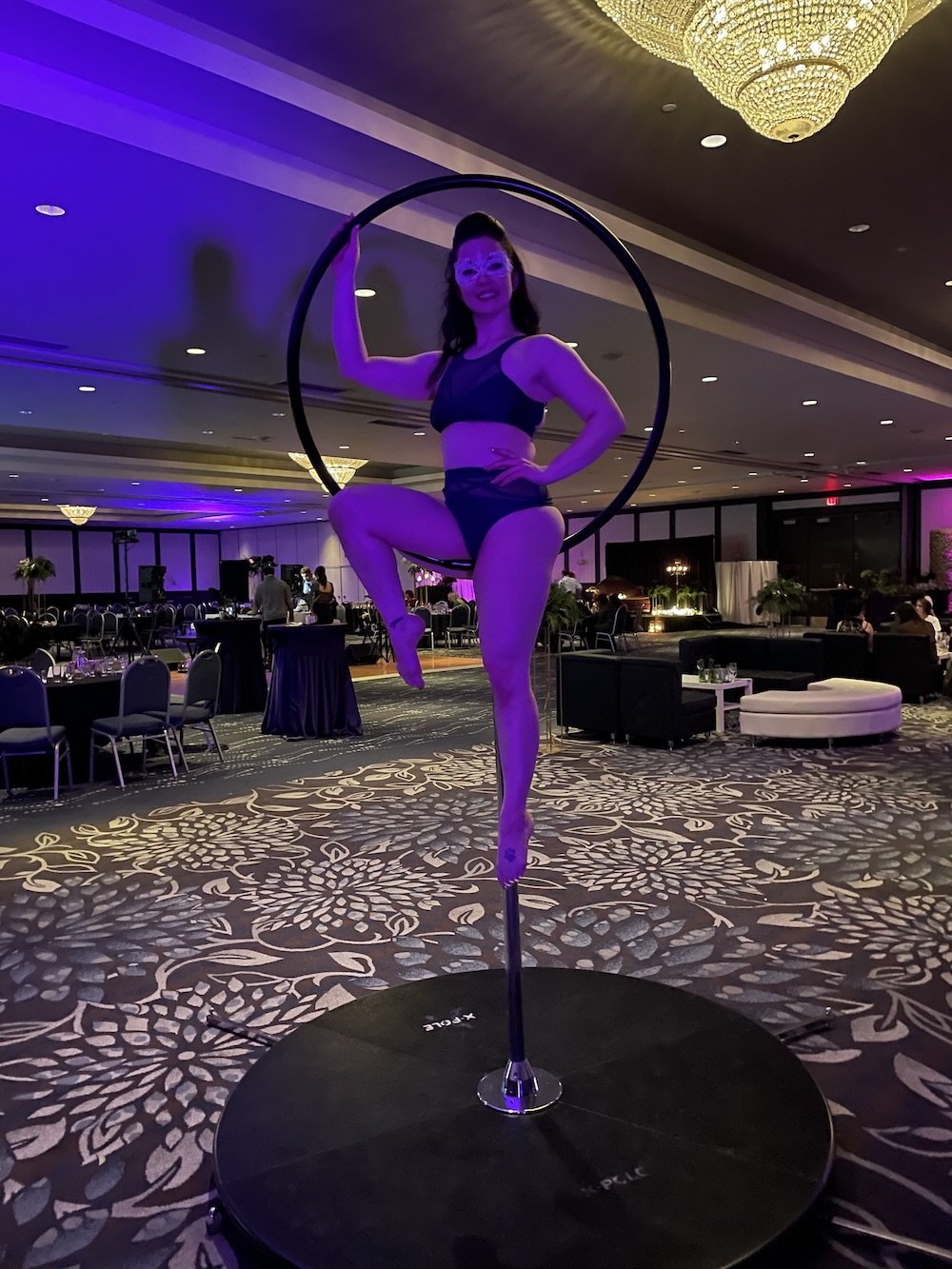 A woman performing a pole dance act on a black pole on a raised platform in a large event hall or ballroom with purple lighting, tables, chairs, and a chandelier.