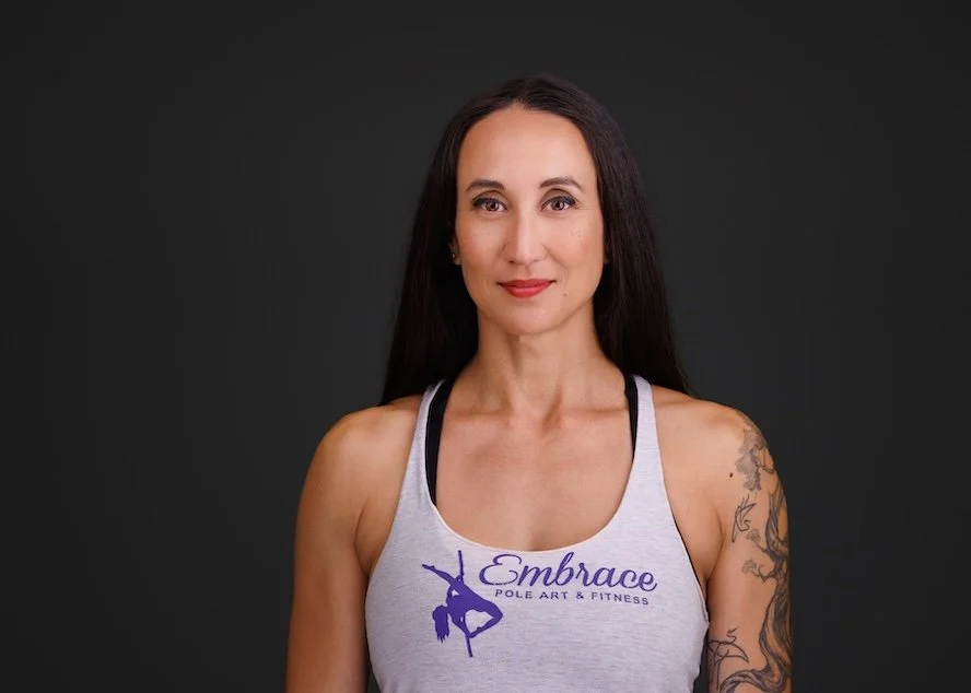 A woman with long dark hair and tattoos on her left arm, wearing a light grey tank top with the logo and text 'Embrace Pole Art & Fitness' against a black background.