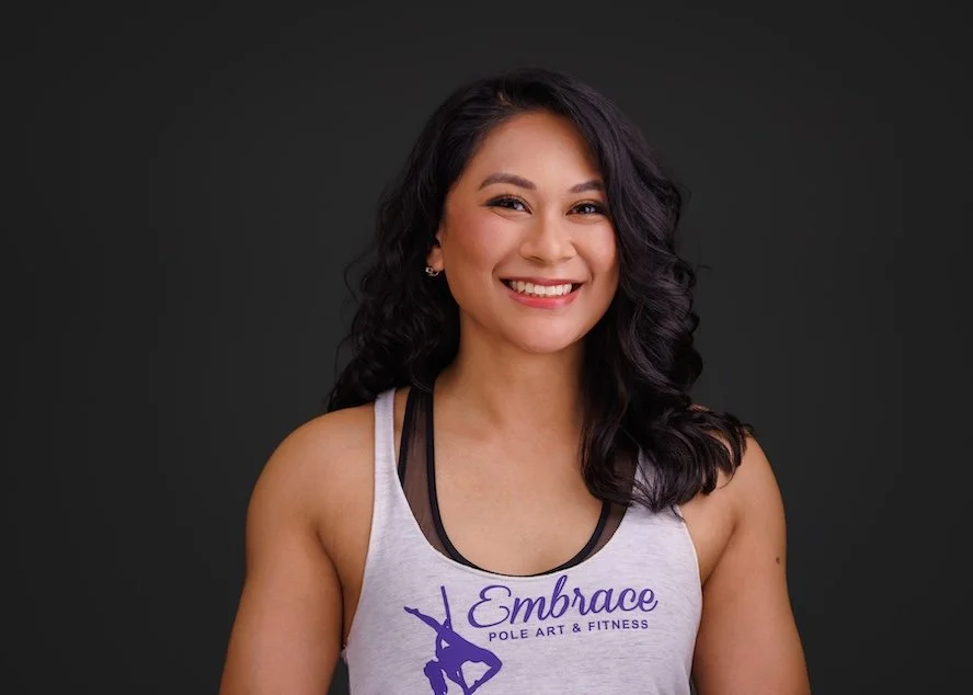 A smiling woman with black, wavy hair wearing a white athletic tank top with purple text that says 'Embrace Pole Art & Fitness' and a silhouette of a pole dancer.