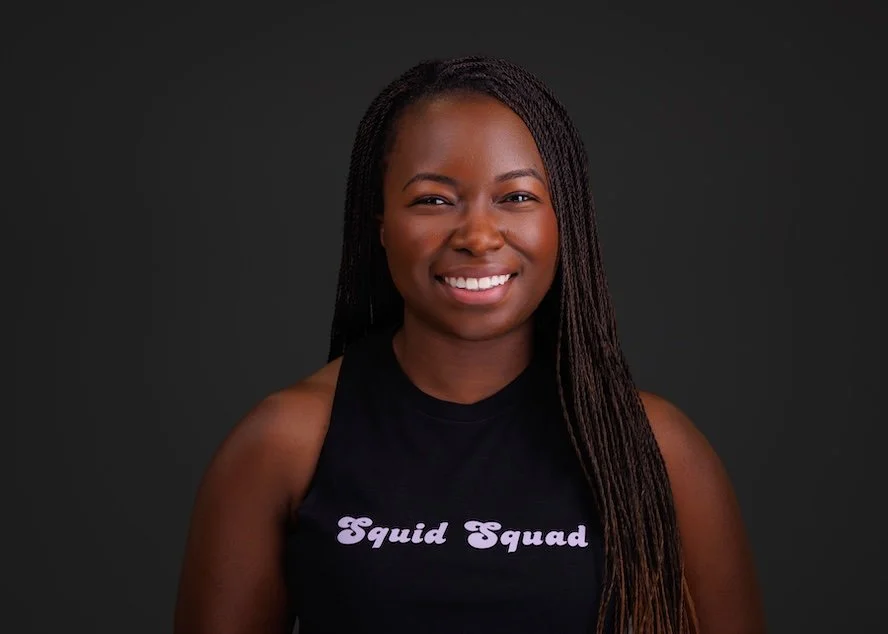 Portrait of a young woman with long braided hair, smiling, wearing a black sleeveless top with 'Squid Squad' written in purple on the front, against a dark background.