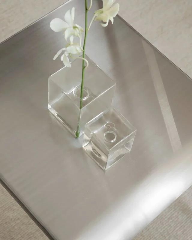 A white flower in a clear square glass vase on a metallic table.