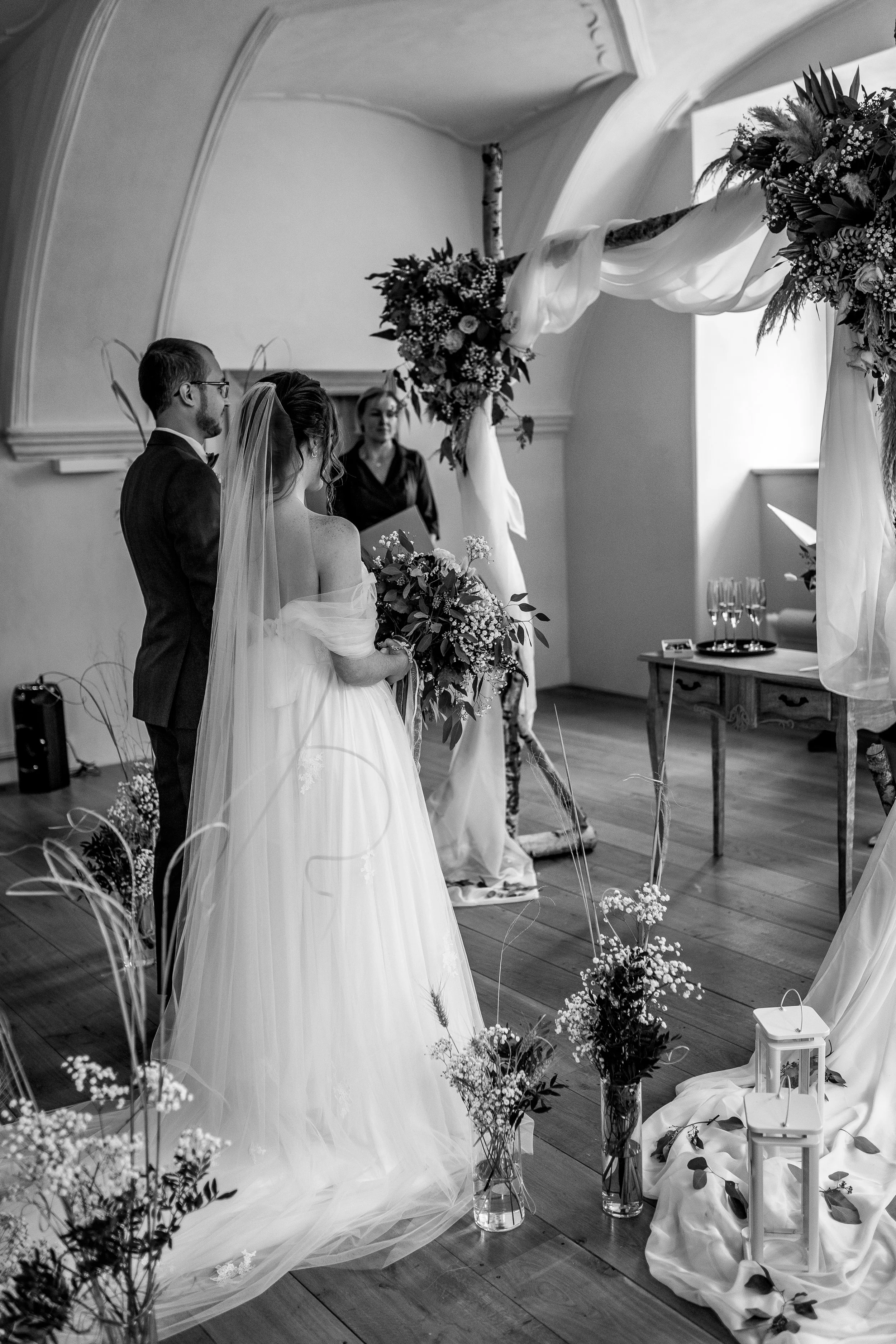 A wedding ceremony with the bride and groom standing before an altar decorated with flowers and draped fabric, in a room with wooden floors and an arched ceiling.