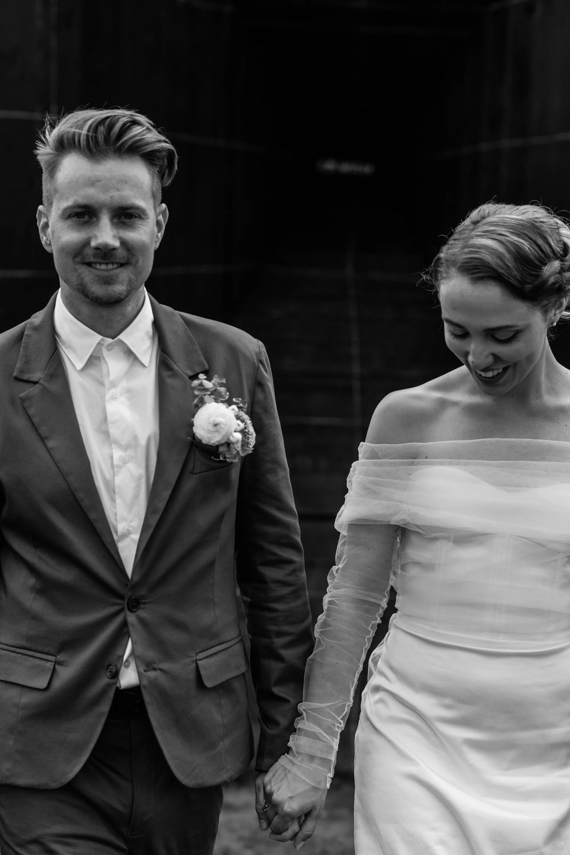 A black and white photo of a man in a suit and a woman in an off-shoulder dress holding hands, smiling, in an indoor setting.