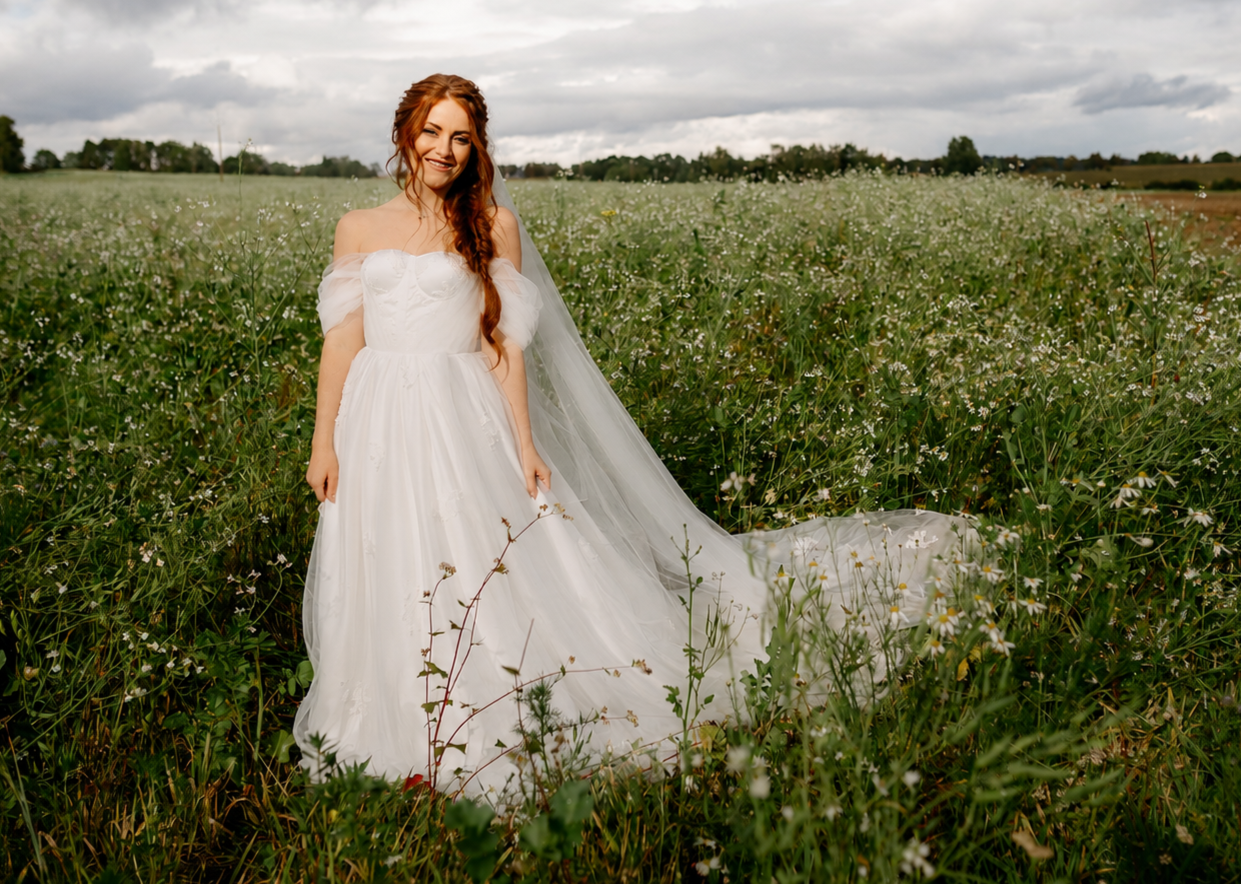 A bride with long red hair in a white wedding dress standing in a field of green plants and small white flowers under a cloudy sky.