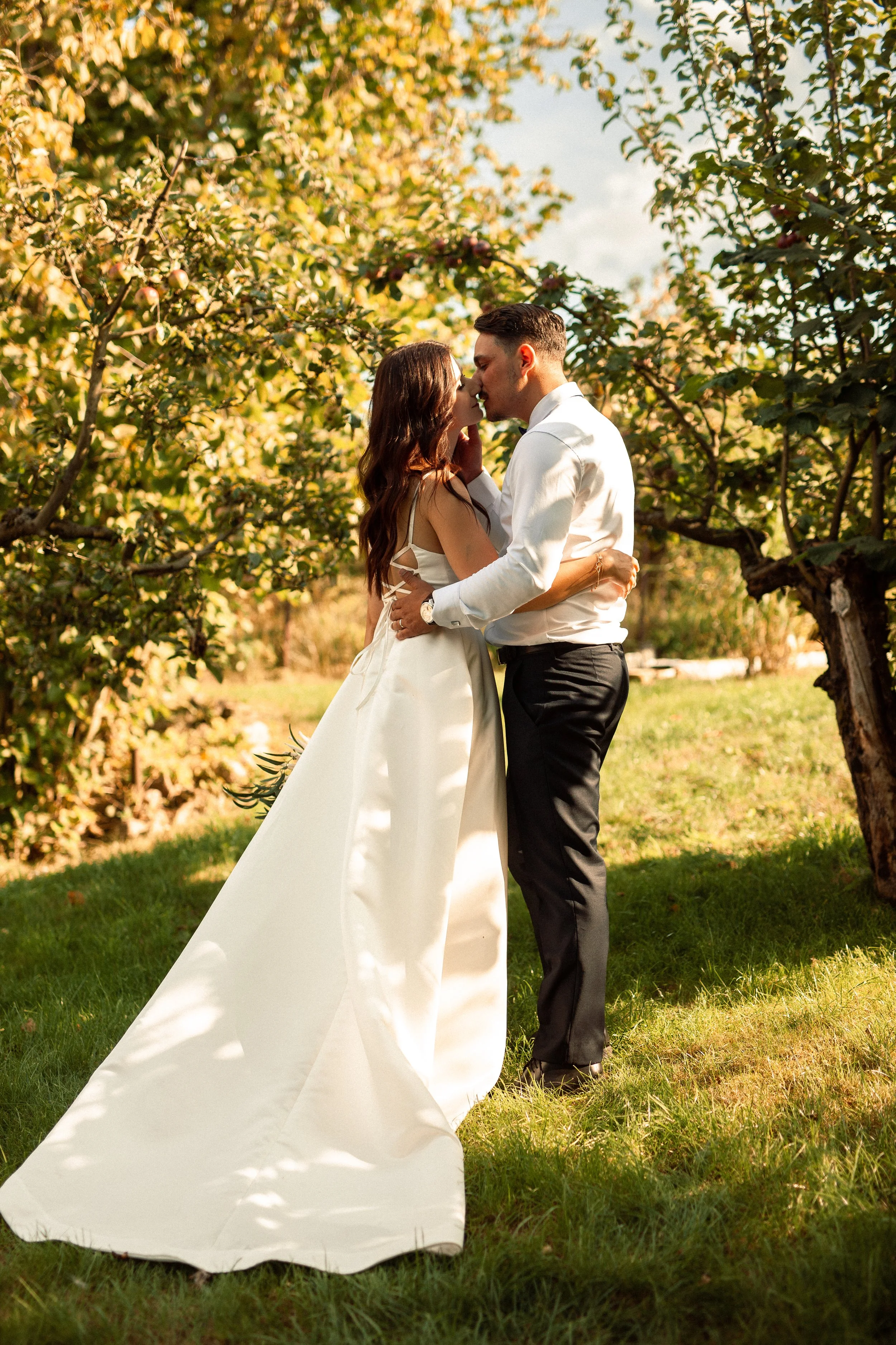 A couple dressed in wedding attire sharing a kiss outdoors among trees with green and yellow leaves, suggesting an autumn setting, during daylight.