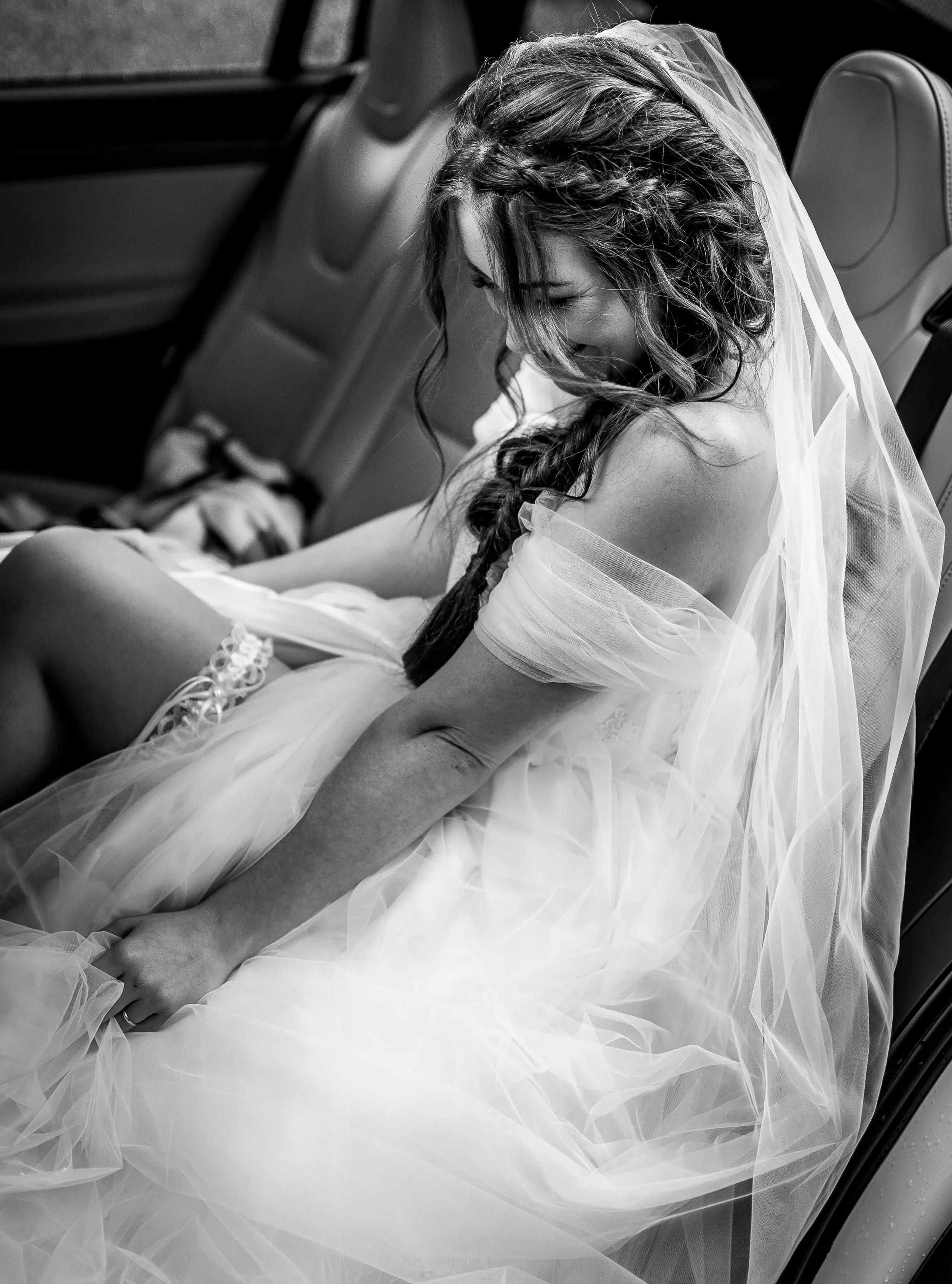 A bride sitting in the backseat of a car, smiling with her eyes closed, wearing a wedding dress and veil.