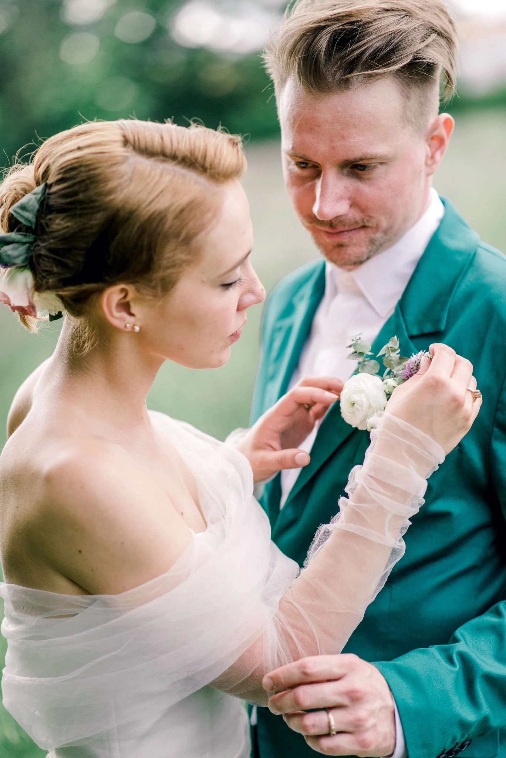 A bride and groom close together, with the bride gently touching the groom's chest, holding a bouquet of flowers, outdoors in a green setting during the daytime.