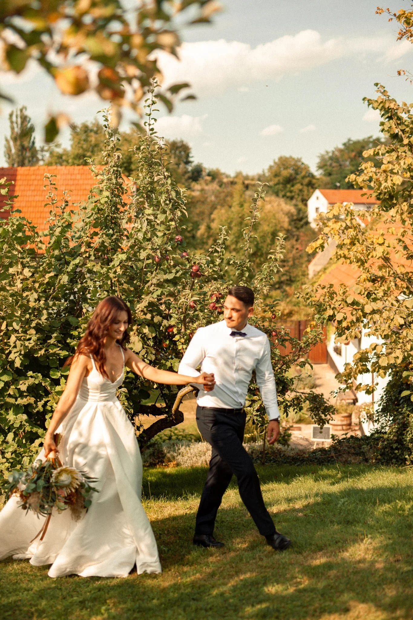 A bride and groom walking hand in hand outdoors in a garden with trees and houses in the background during daytime.